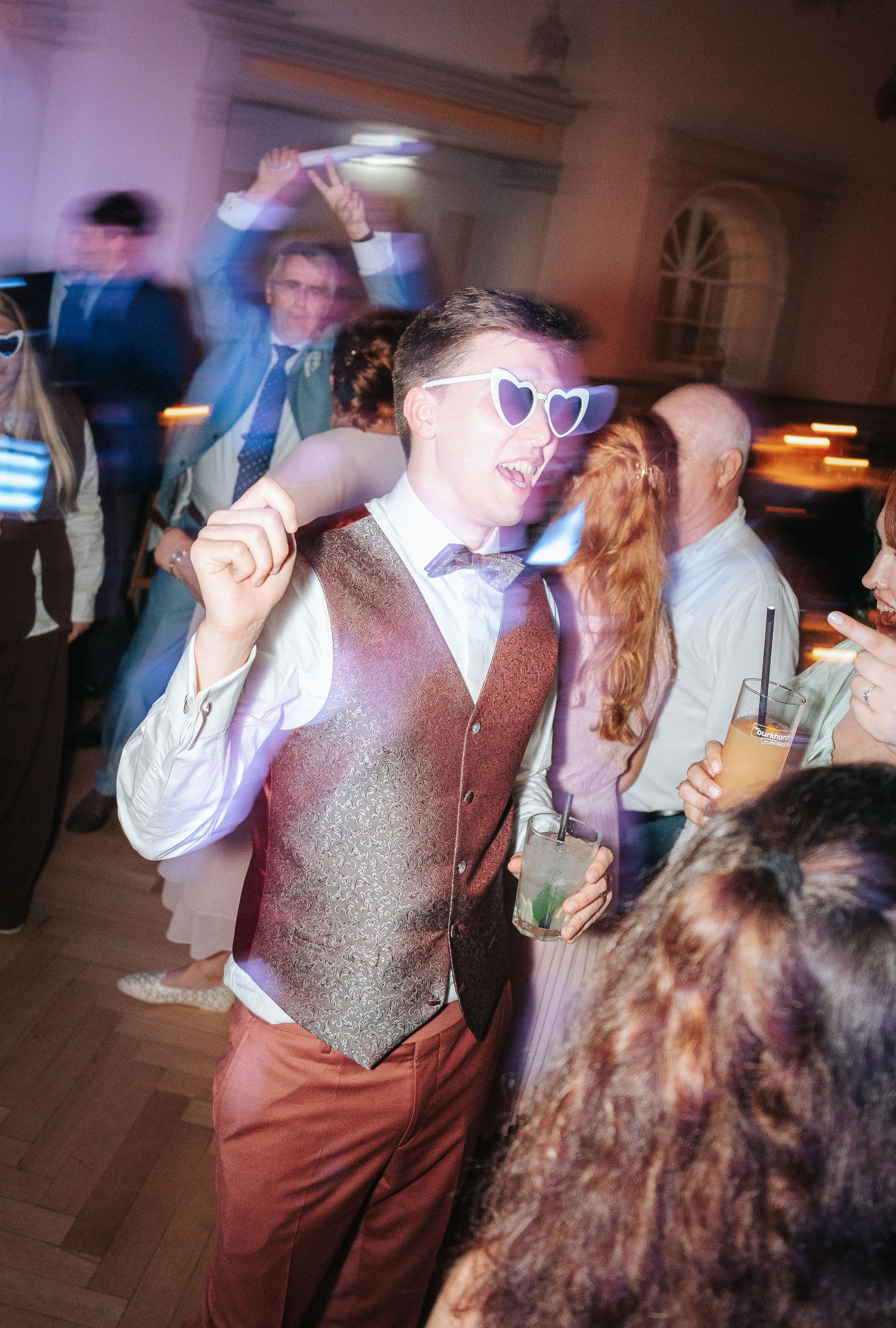 Man in heart-shaped sunglasses, glittery vest and bow tie dances with a drink at a lively wedding reception.