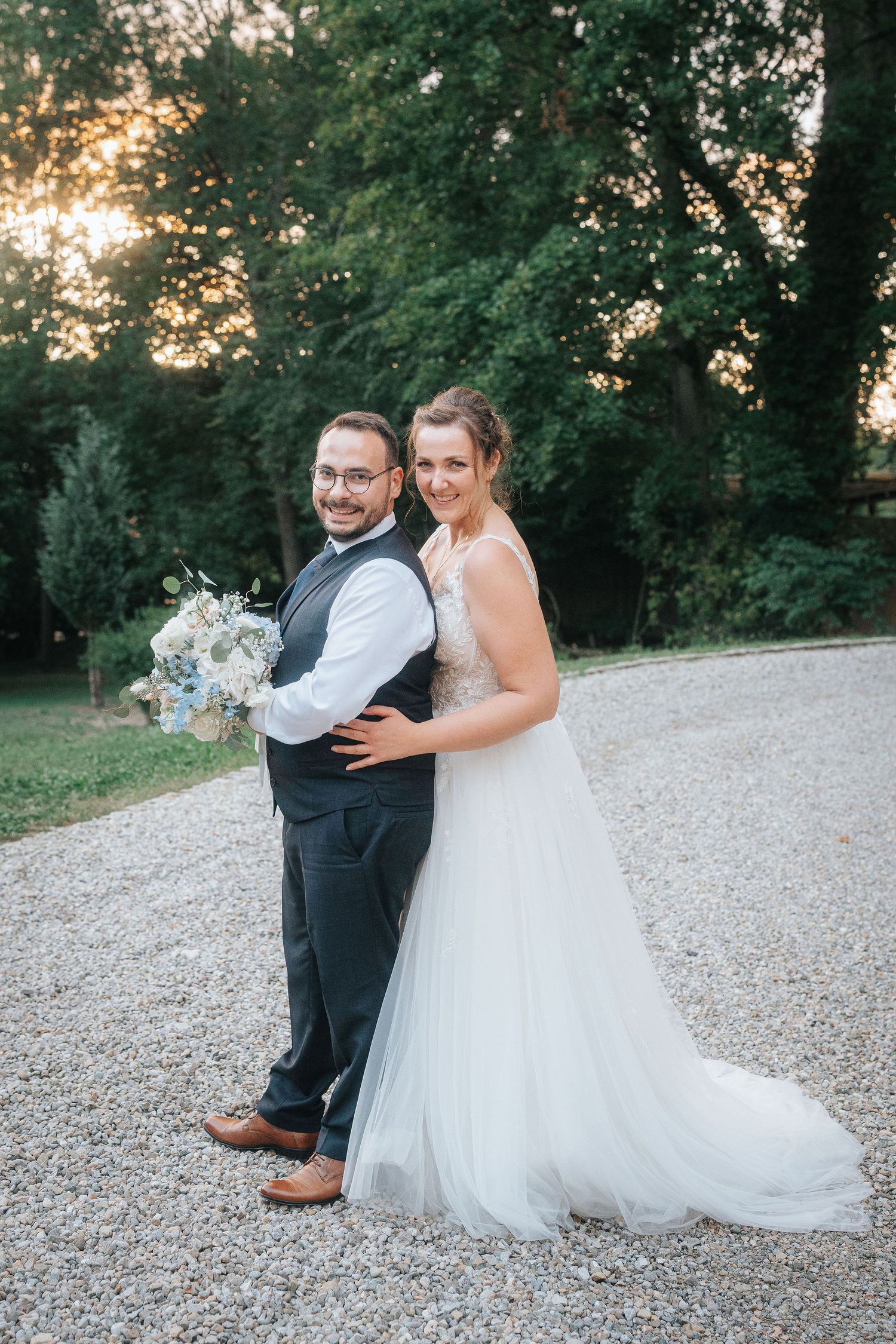 Smiling bride in a white tulle gown embraces a groom in a vest holding a blue bouquet on a gravel path with trees at sunset.