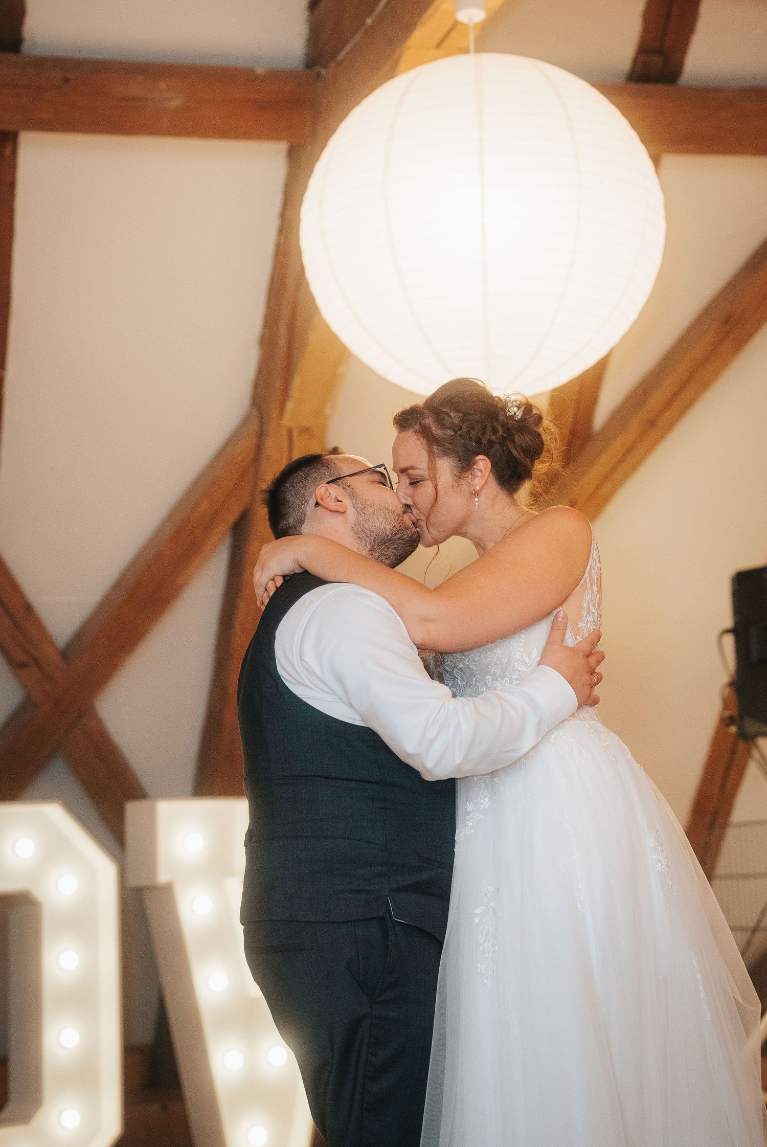 Bride in a white tulle gown and groom in a dark vest share a kiss beneath a paper lantern and wooden beams, with light-up letters nearby.