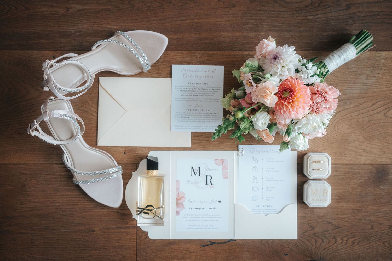 Flat lay of wedding details on wood with a pink peach bouquet, silver strappy heels, perfume bottle, ring boxes and invitation suite.