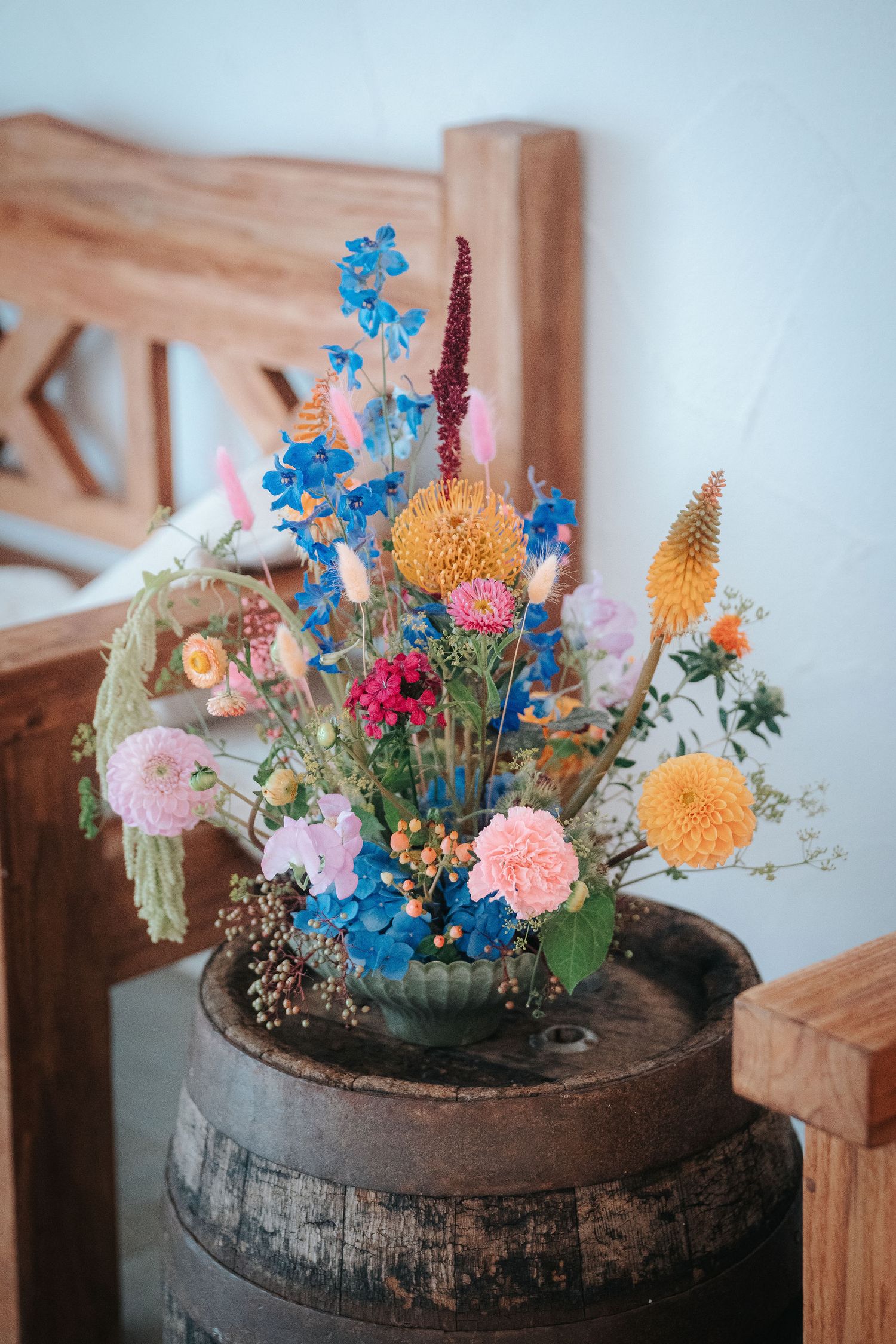 Colorful bouquet of blue delphinium, pink dahlias, and yellow billy buttons in a vase on a rustic wooden barrel beside a carved chair.