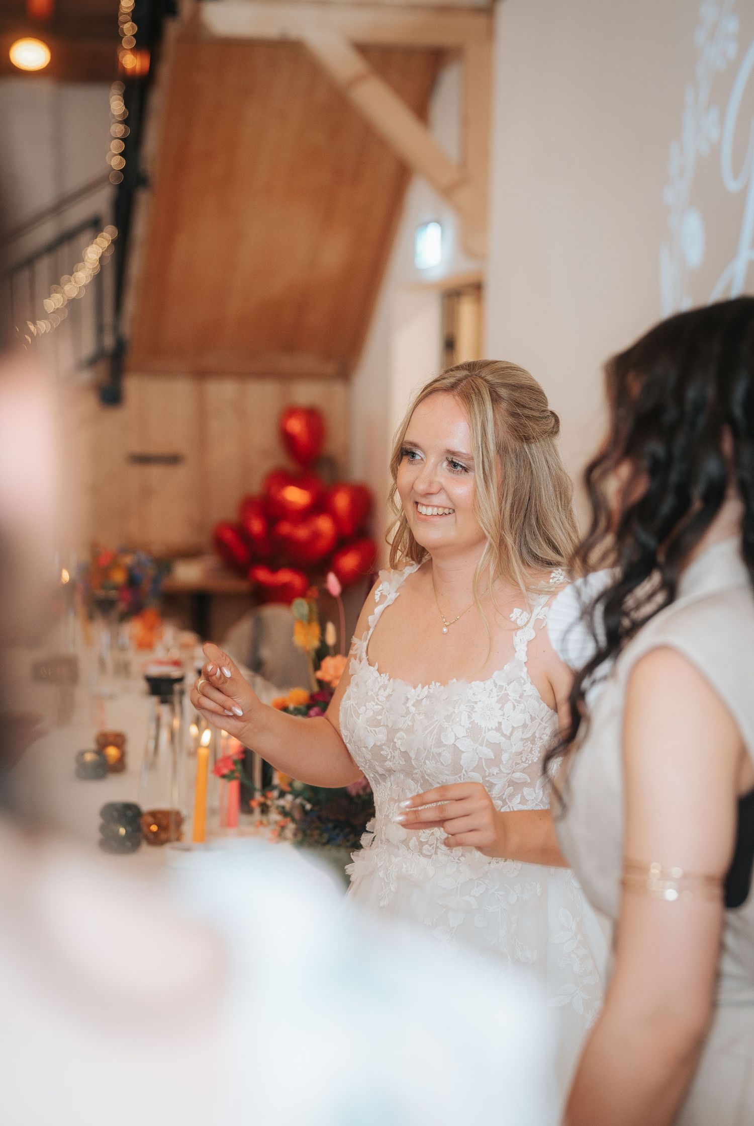 Smiling bride in a lace wedding gown with a braided half-up hairstyle stands at a reception with red heart balloons and candlelit tables.