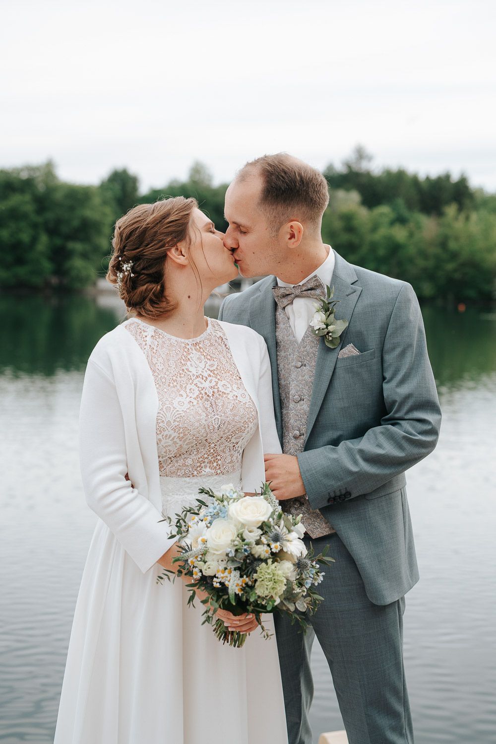 A romantic wedding couple kiss by a serene lake while the bride holds a beautiful white floral bouquet.