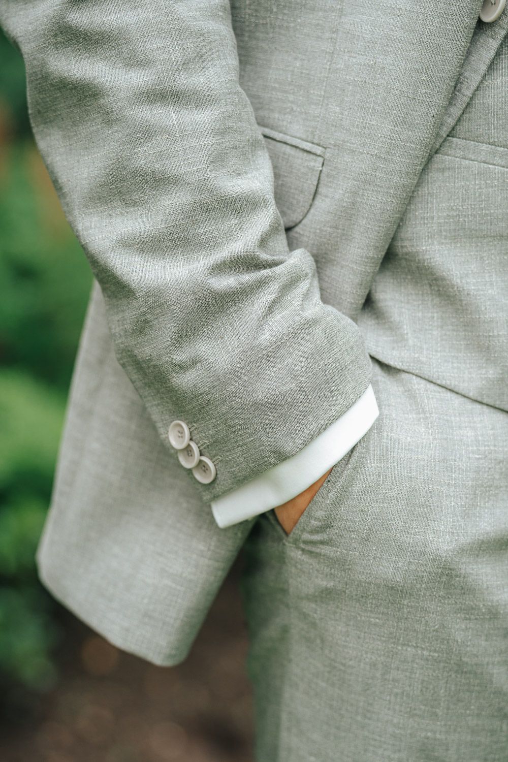 Close-up detail of a light gray suit sleeve with buttons and pocket, showing tailored menswear styling against blurred green background.