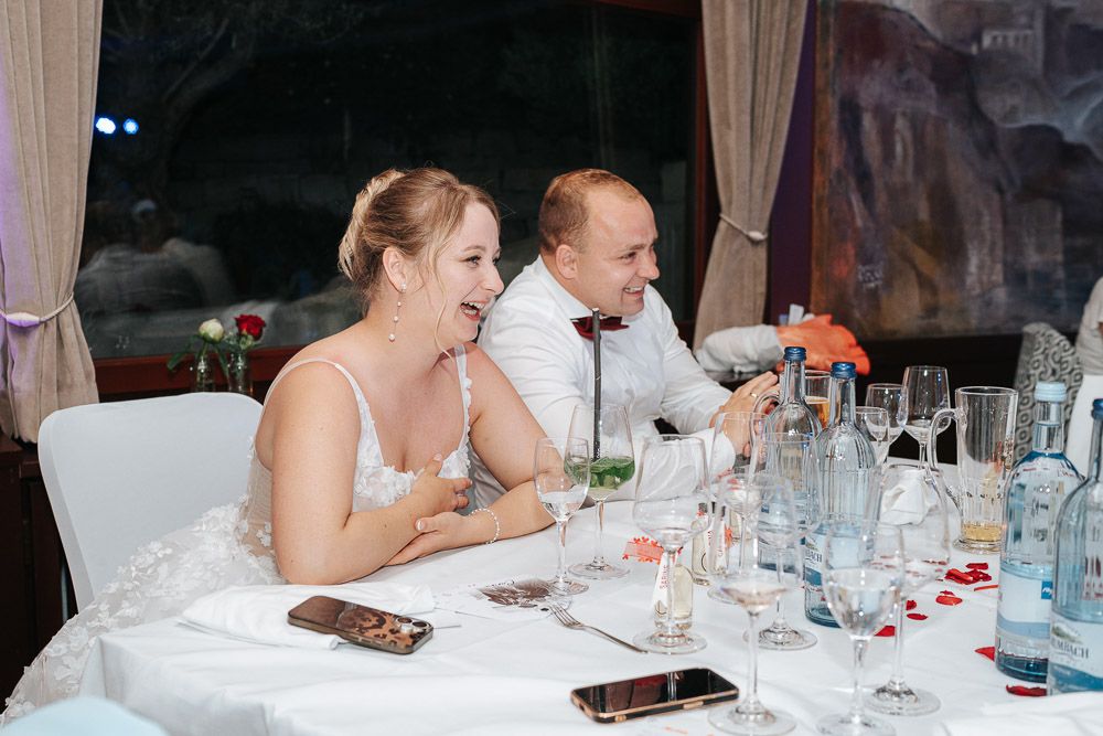 A wedding party sits at a decorated table with glassware and floral centerpieces for a celebratory reception dinner.