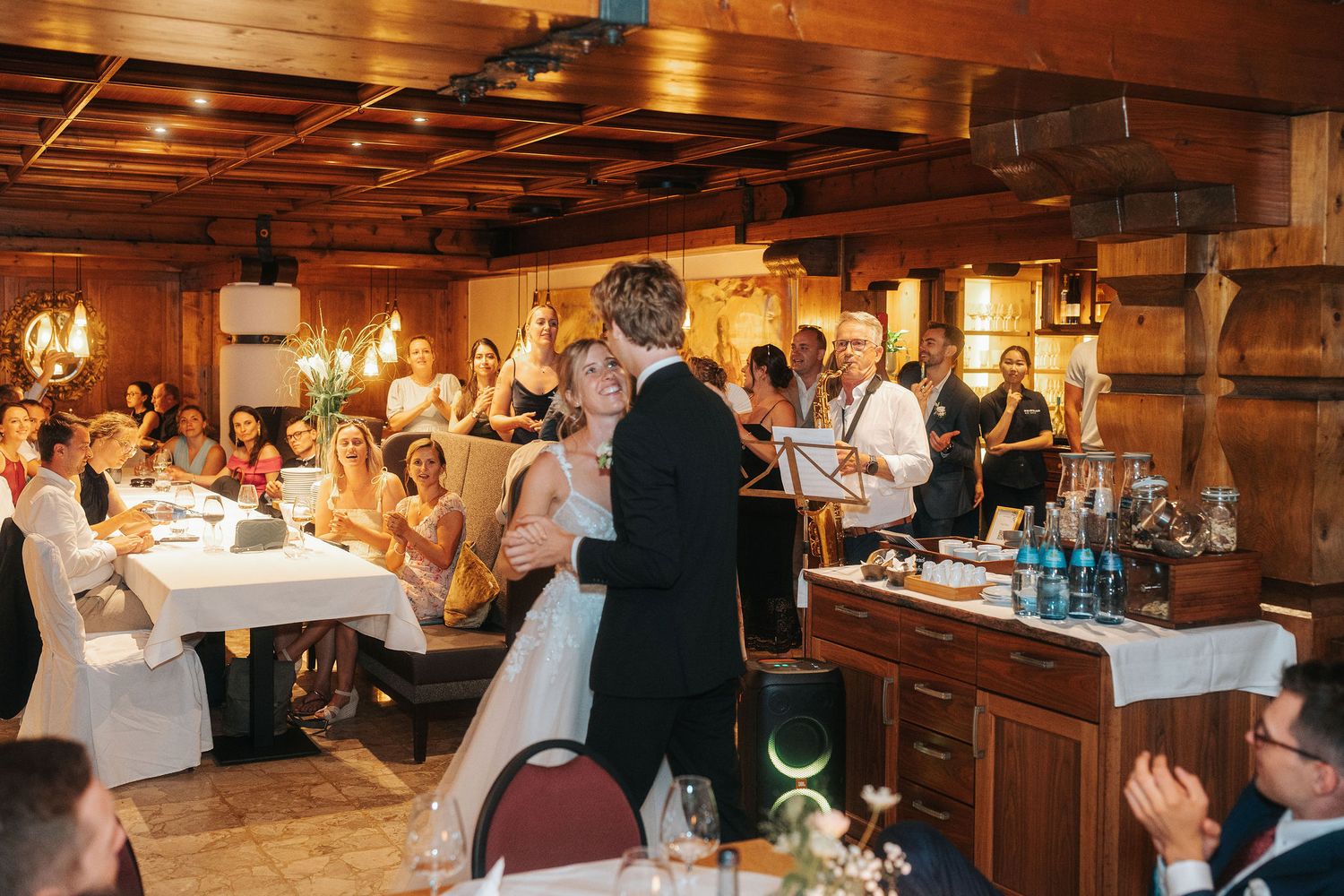 Bride and groom share a first dance in a rustic wooden banquet hall while seated wedding guests watch from tables.
