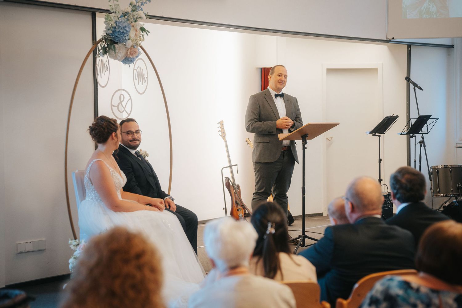A bride and groom sit before wedding guests as an officiant in a gray suit speaks during an indoor ceremony beside a floral hoop.