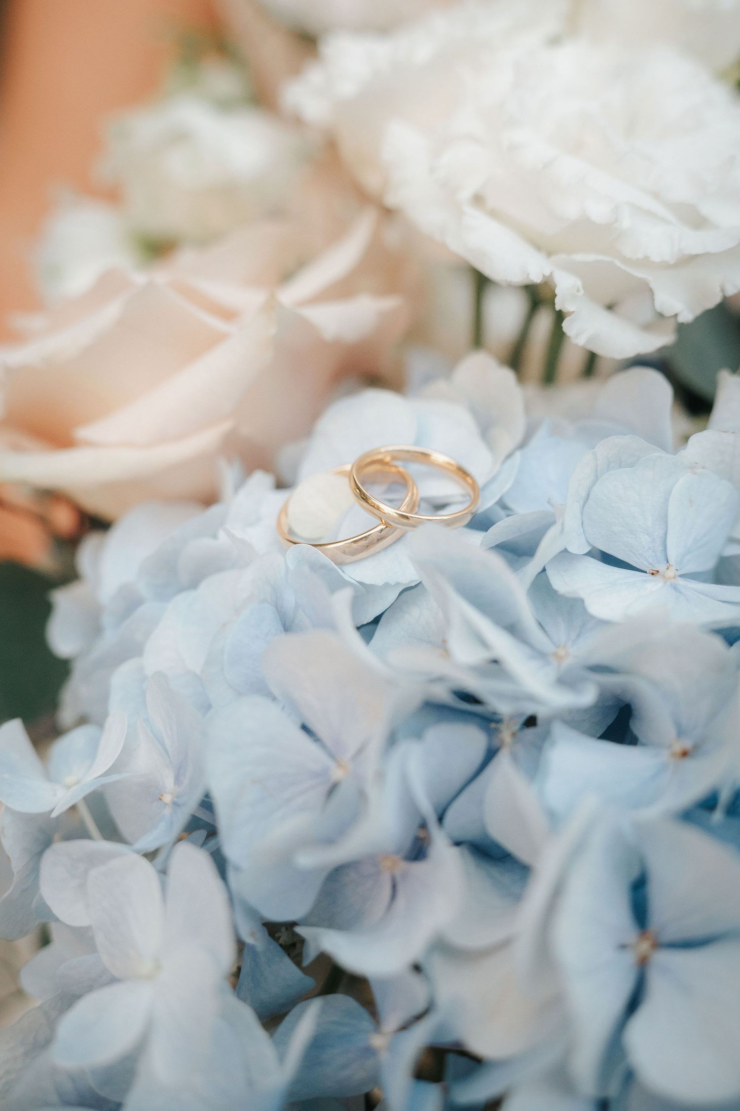 Two gold wedding bands resting on soft blue hydrangea petals with white and blush pink roses in a romantic bridal bouquet.