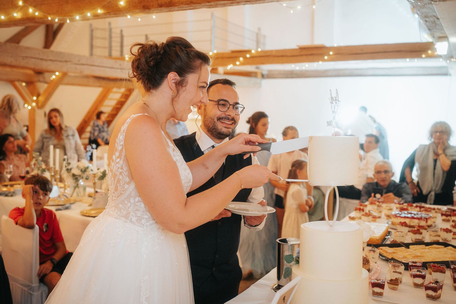 Bride in a white gown and groom in a dark suit smile while cutting their wedding cake together as guests watch in a rustic reception hall.