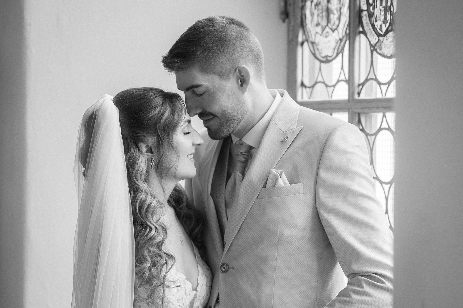 Black and white photo of a bride in a long veil and a groom in a light suit touching foreheads in a tender wedding moment by a window.
