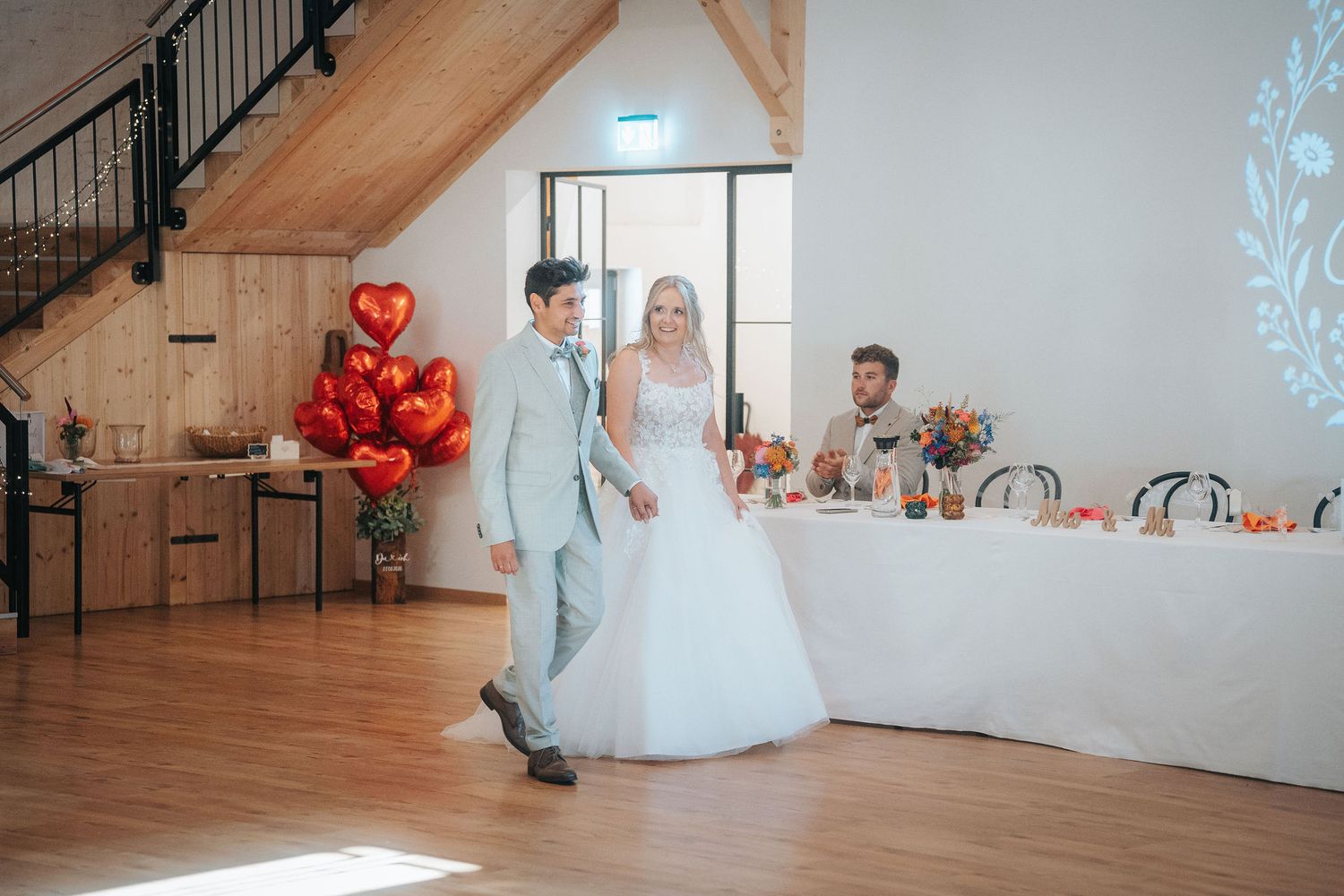 Bride in a white gown and groom in a light gray suit walk hand in hand across a wood floor past red heart balloons at a rustic wedding recep