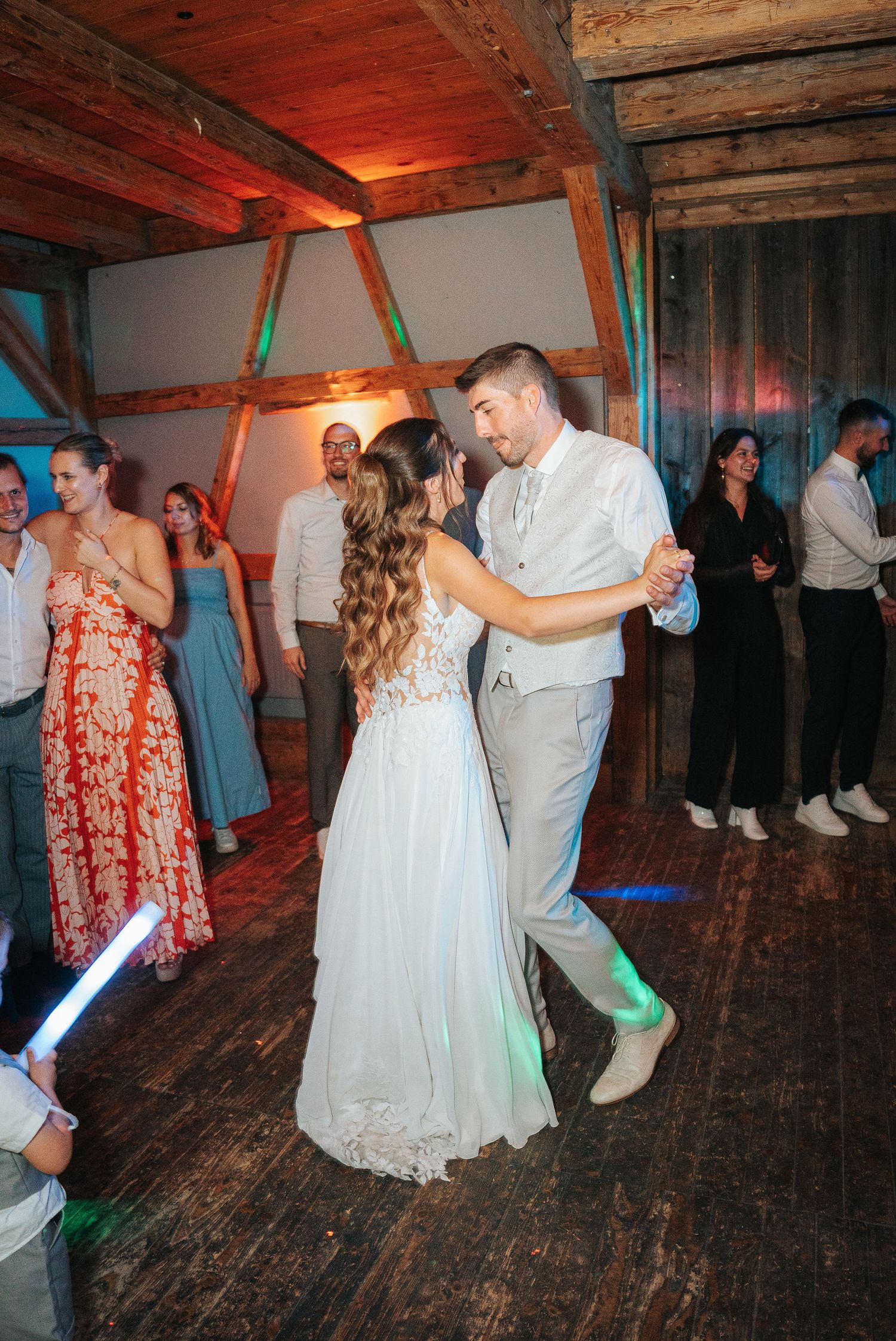 Bride in a lace wedding gown and groom in a gray vest dance as smiling guests watch in a rustic timber-framed venue.