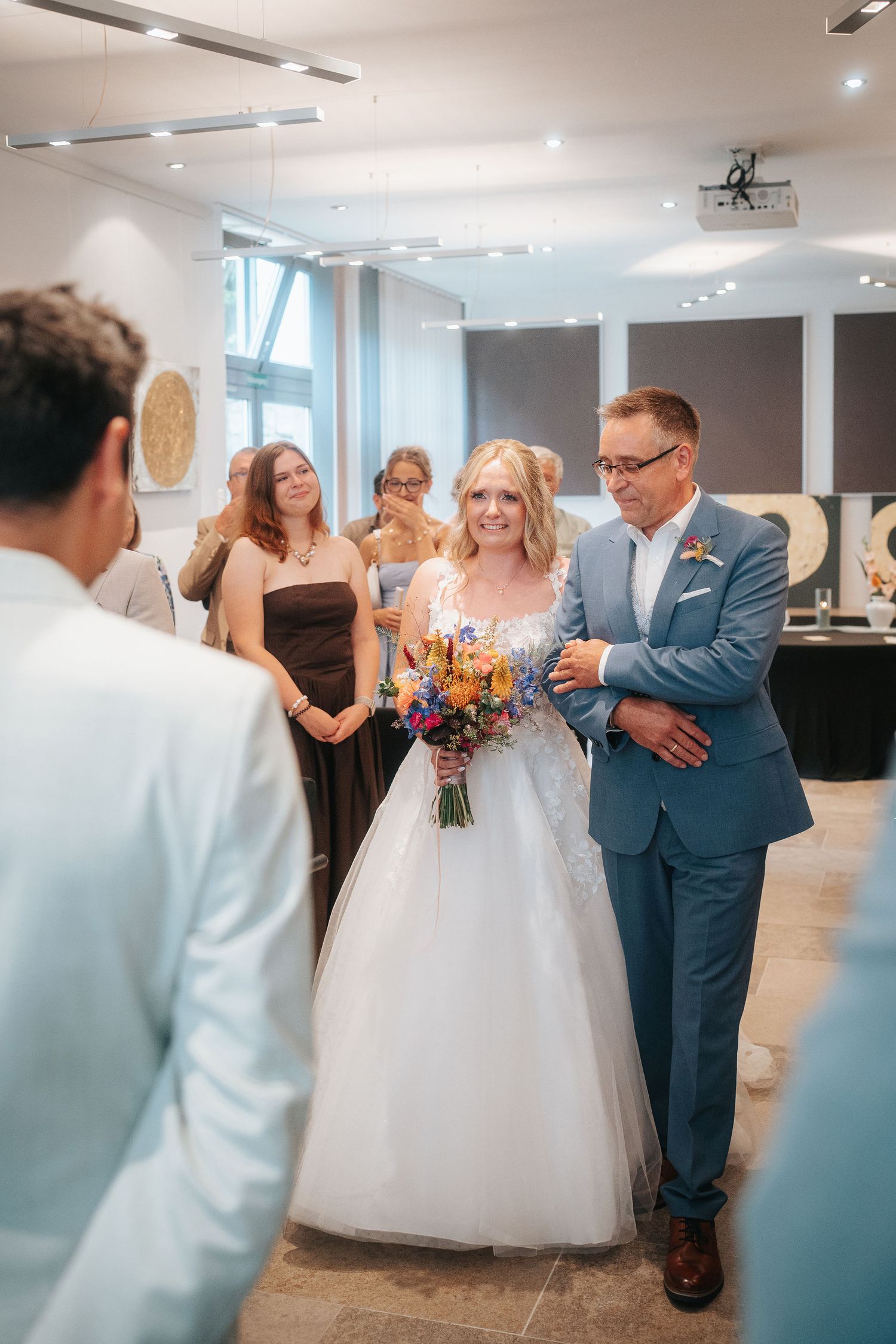 Smiling bride in a white tulle gown holding a colorful bouquet walks down the aisle on her father's arm as wedding guests look on.