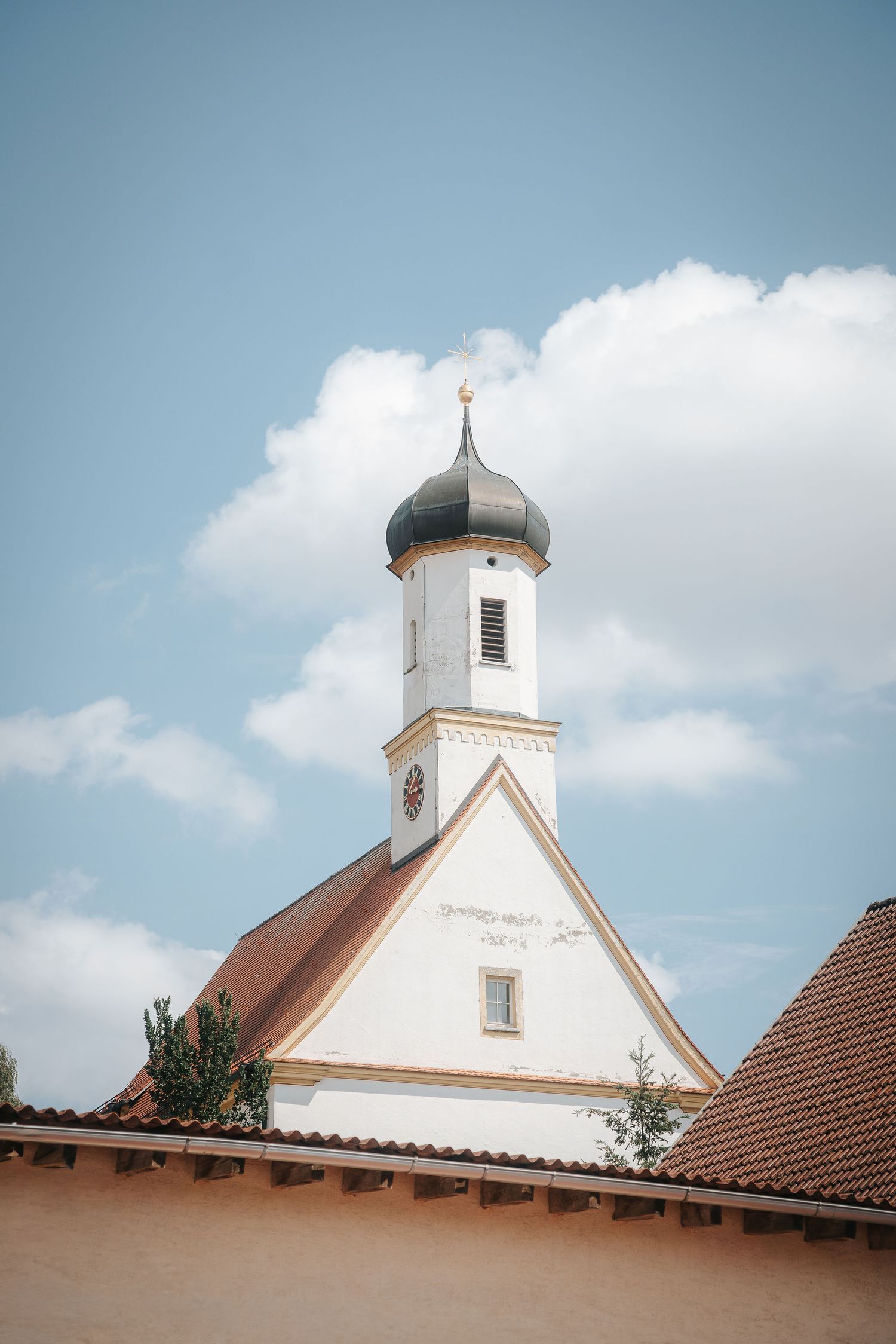 White church with an onion dome tower, clock and red tile roof rising above a rooftop against a blue sky with clouds.