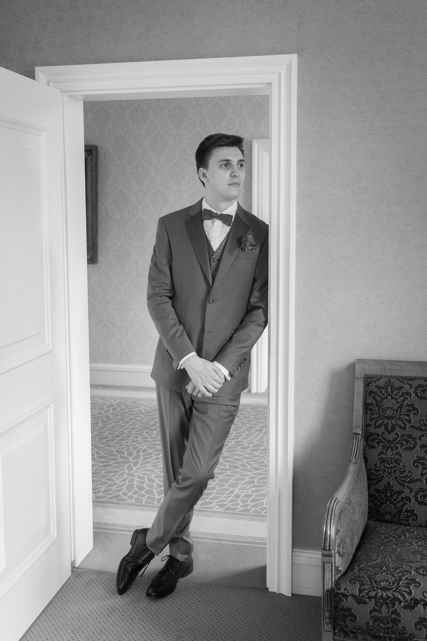 Black and white photo of a groom in a suit, vest and bow tie leaning casually against a doorframe in an elegant room.