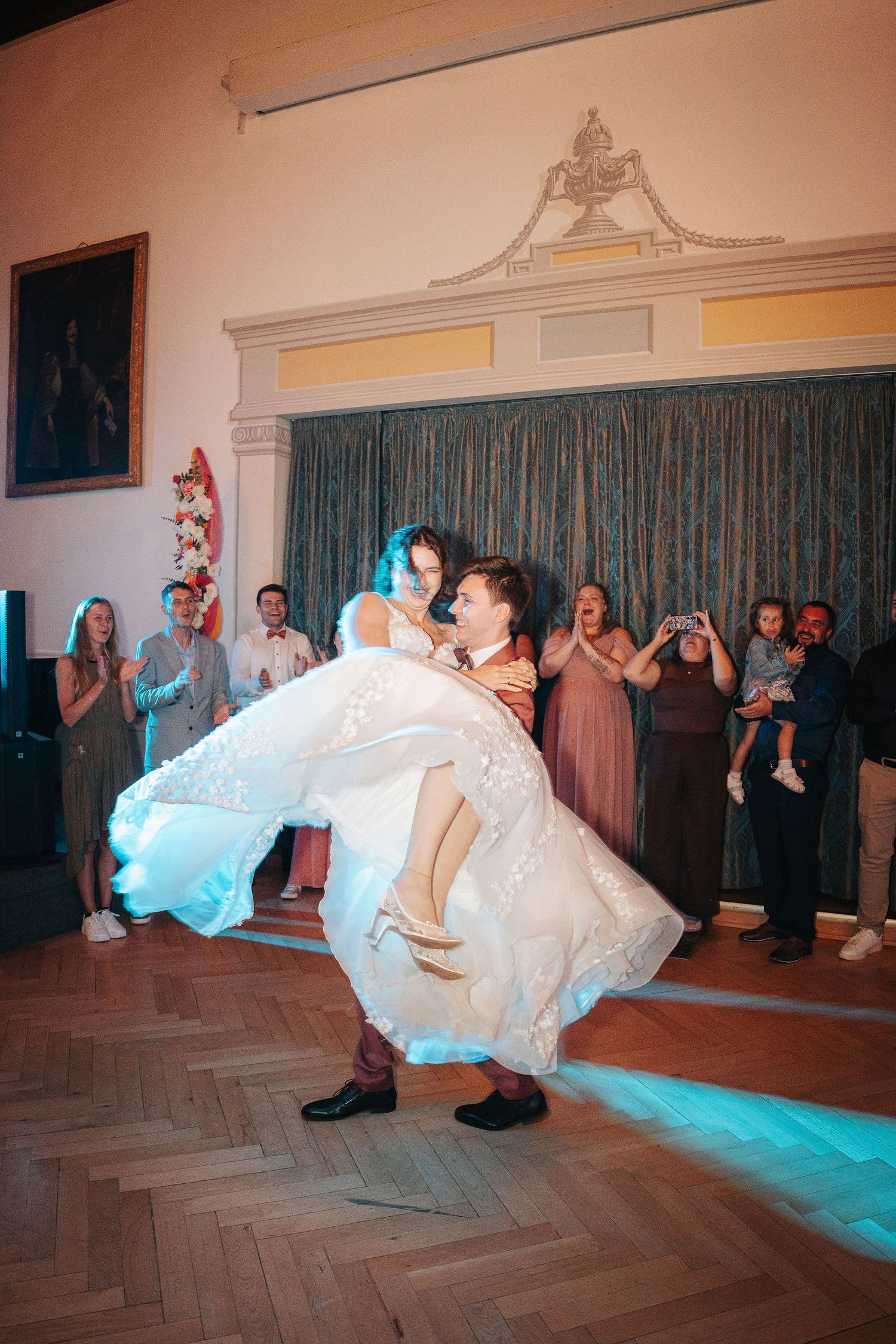 Groom lifts bride in a flowing white wedding gown during their first dance as smiling guests watch in an elegant ballroom.