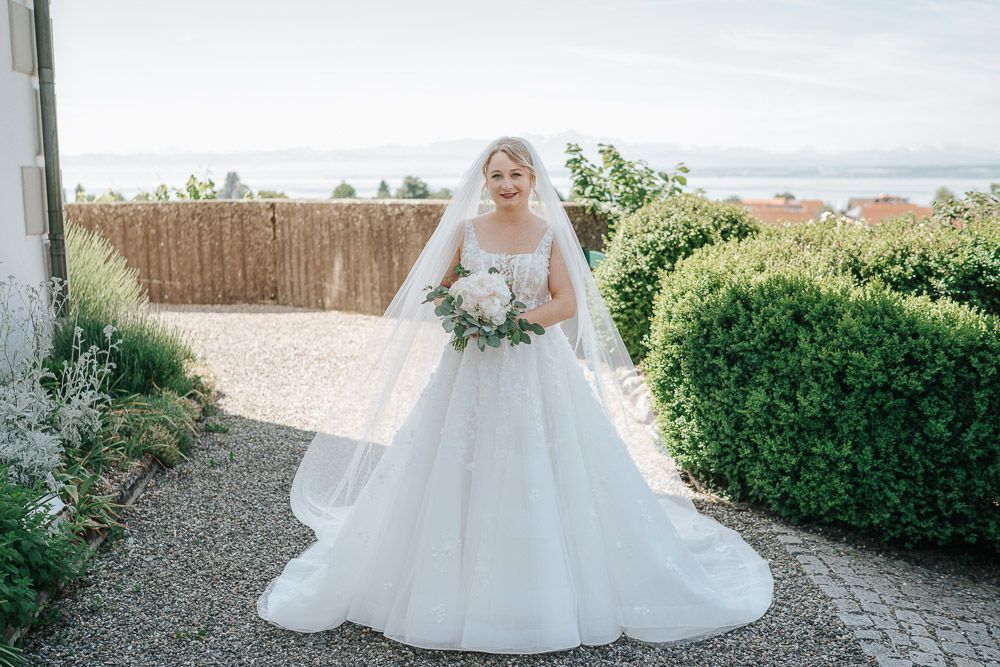 A bride in a white ballgown wedding dress and long veil stands on a gravel path overlooking a scenic coastal view.