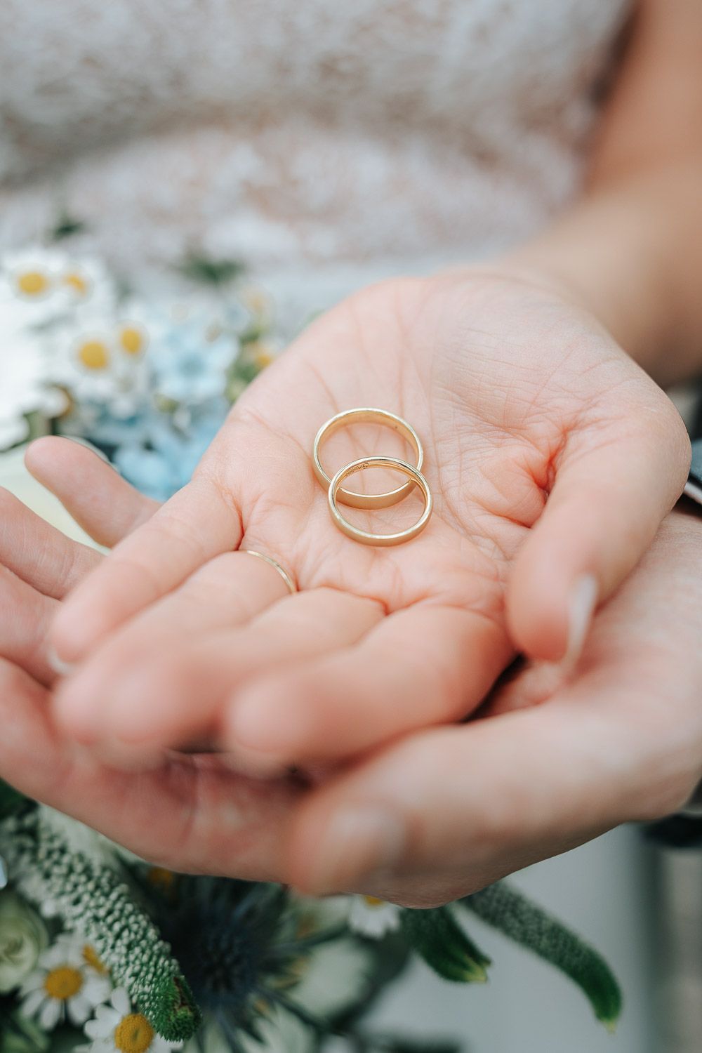 Two gold wedding rings held gently in open palms with white daisies and greenery in the background.