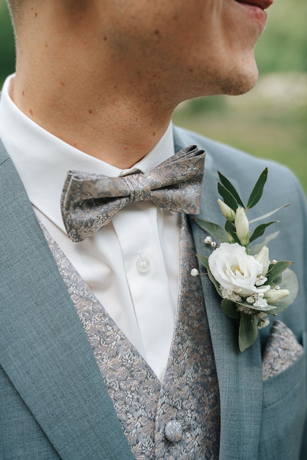 A groom's boutonniere features a white rose and greenery against a sage green suit for an elegant wedding look.