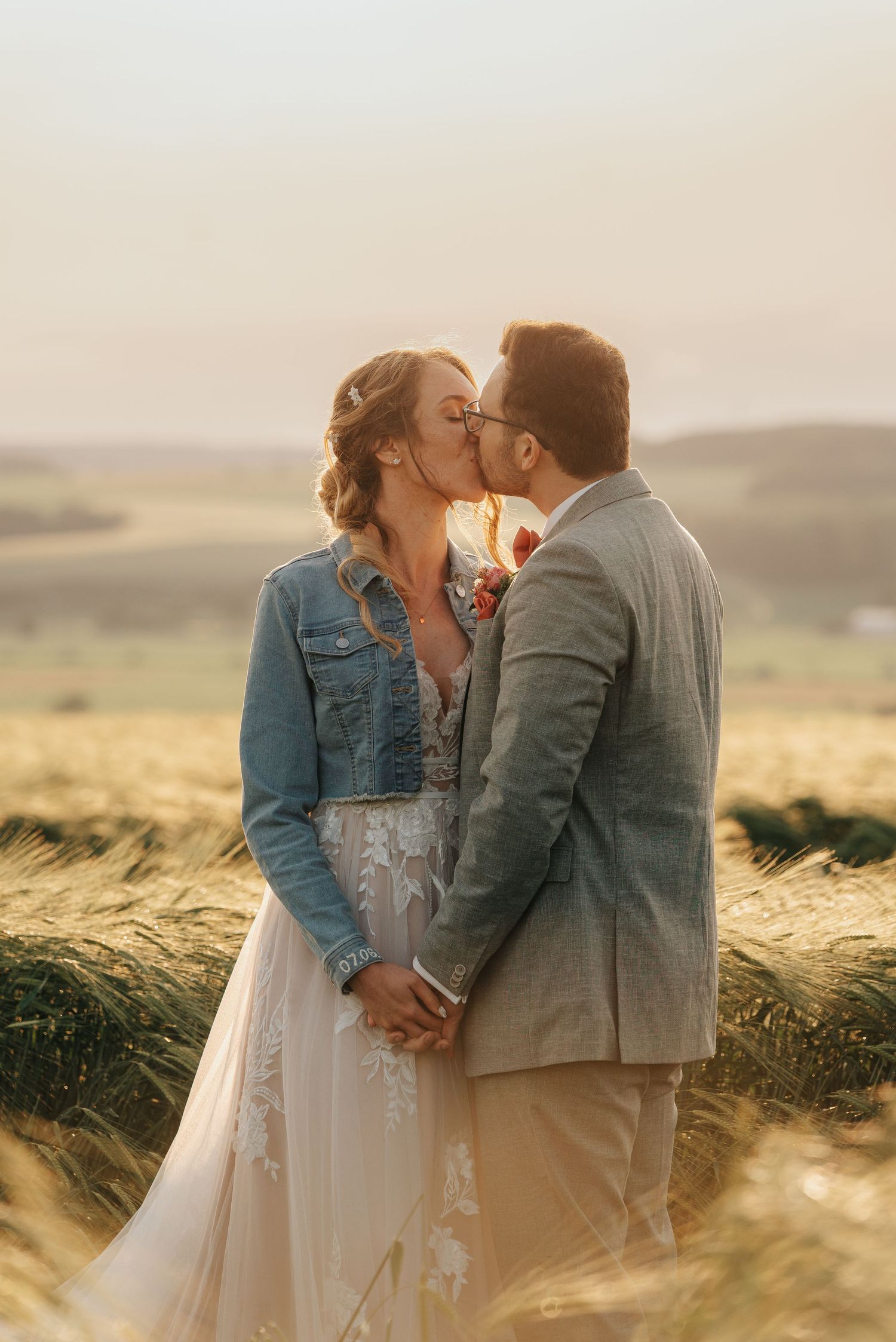 Couple shares romantic kiss in golden wheat field at sunset wearing denim jacket and gray suit.