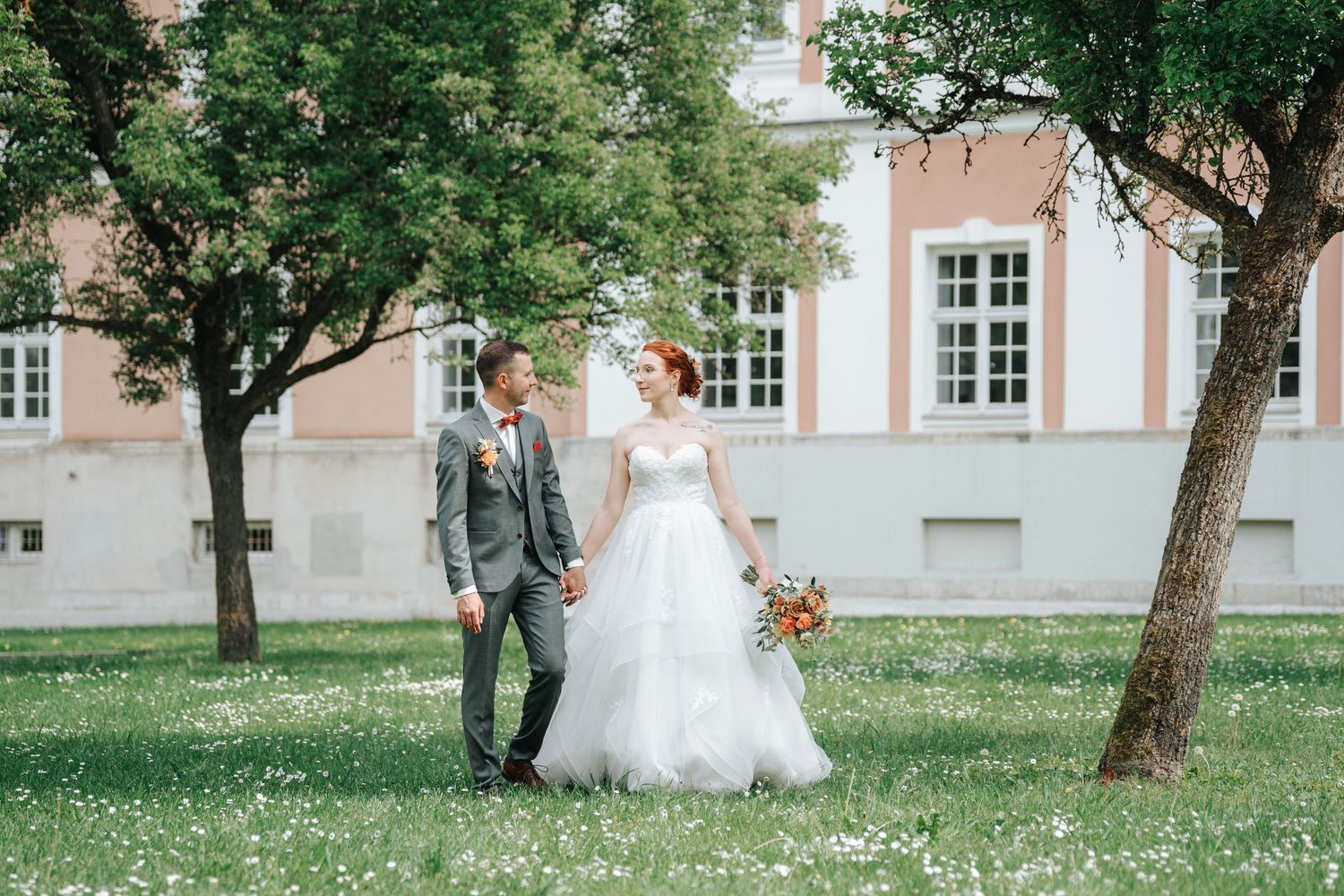 Bride and groom walking hand in hand on a flower-filled lawn beside a pink historic building on their wedding day.
