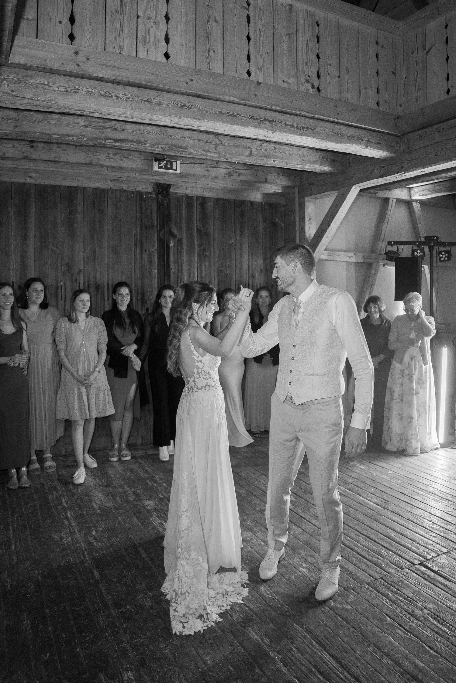 Bride in a lace gown and groom in a vest share a first dance on a wooden barn floor as wedding guests watch in black and white.