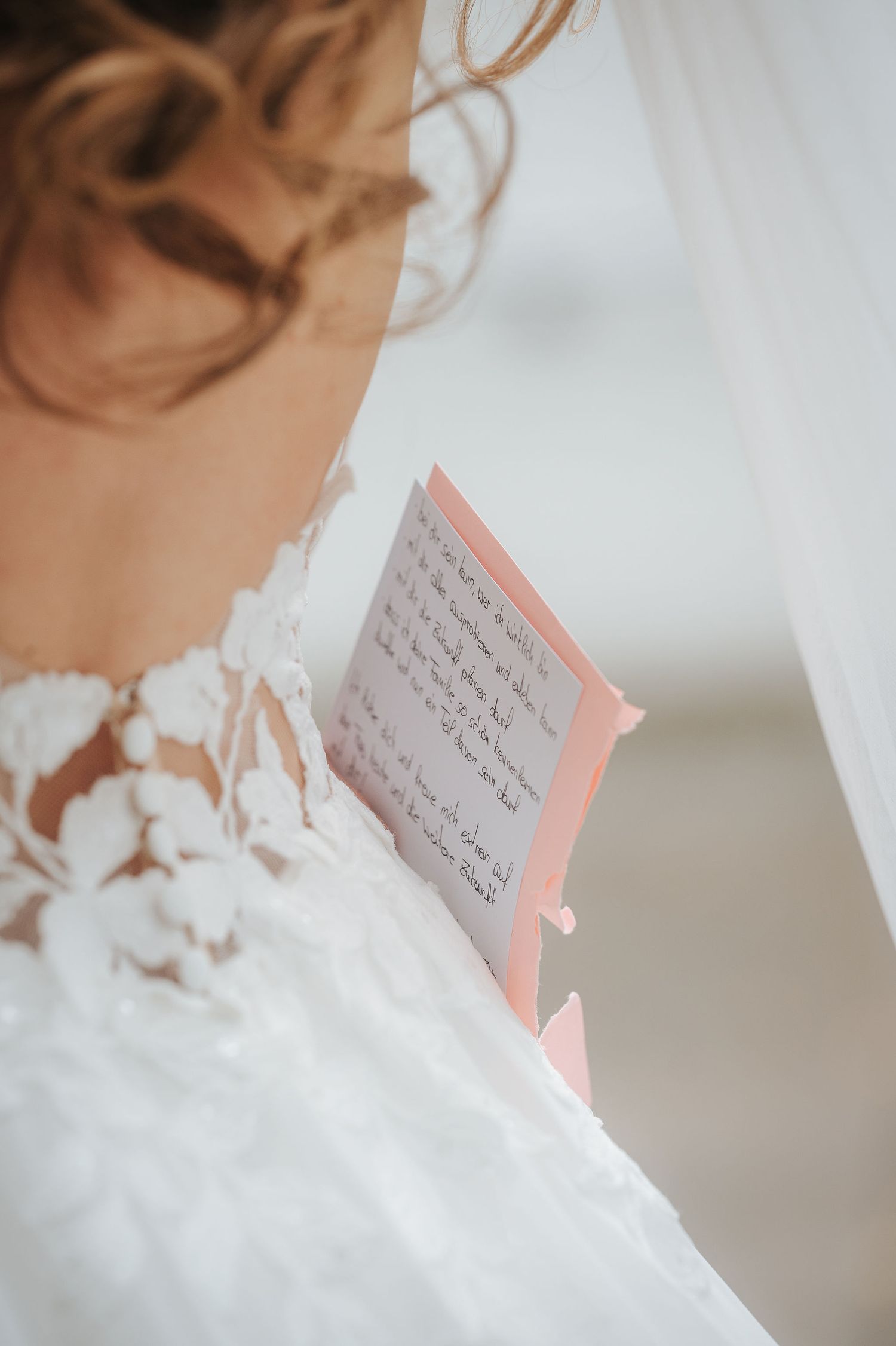 Bride in a white lace wedding gown with an open back reads handwritten vows from a pink notecard during an outdoor ceremony.