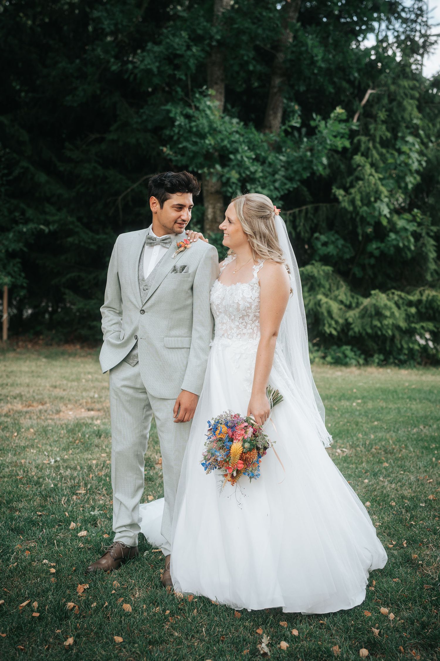 Smiling groom in a light gray suit and bow tie embraces a bride in a lace gown and veil holding a colorful bouquet on a grassy lawn.