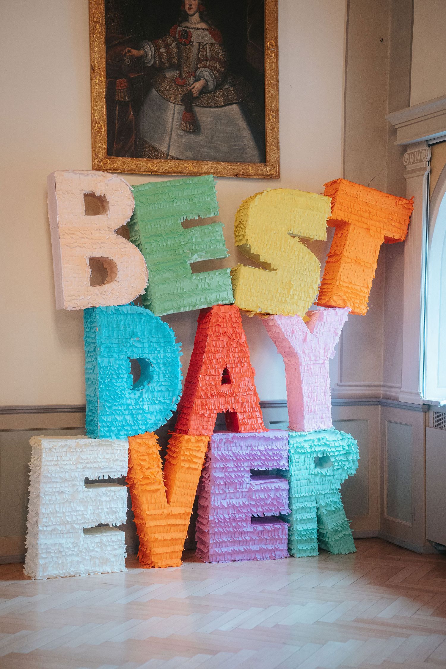 Colorful fringed piñata letters spelling BEST DAY EVER stacked on a wooden floor beneath a framed vintage portrait.