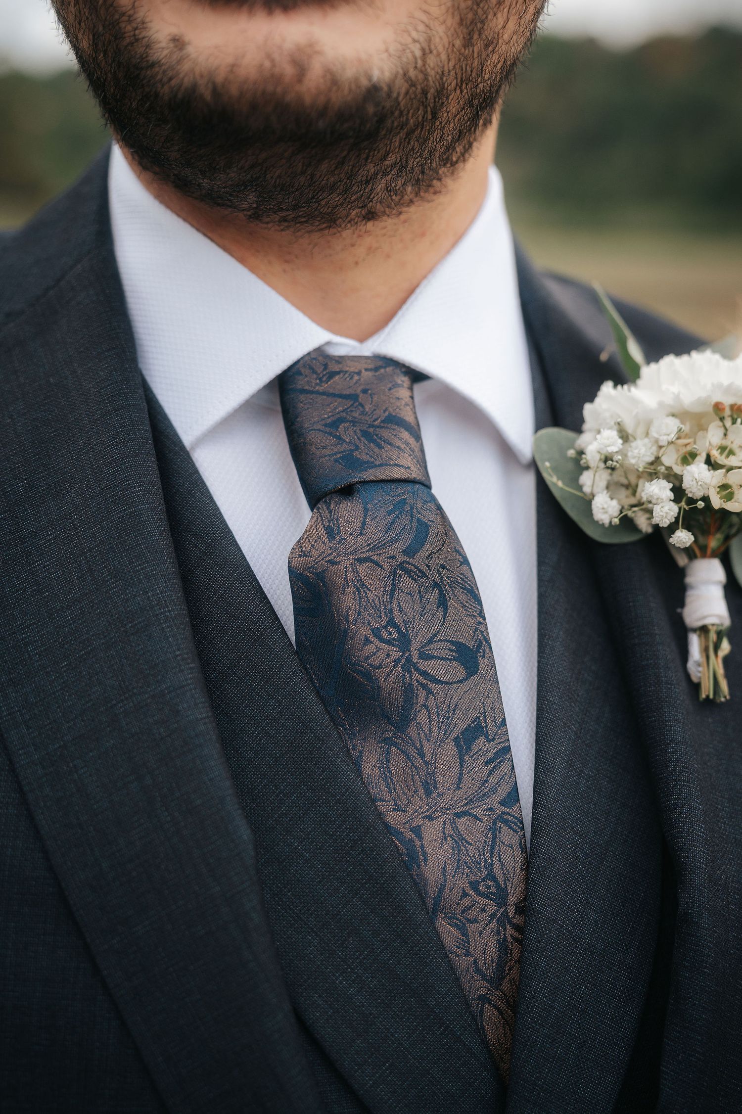 Close-up of a groom in a charcoal suit with a patterned navy and bronze floral tie and a white baby's breath boutonniere.