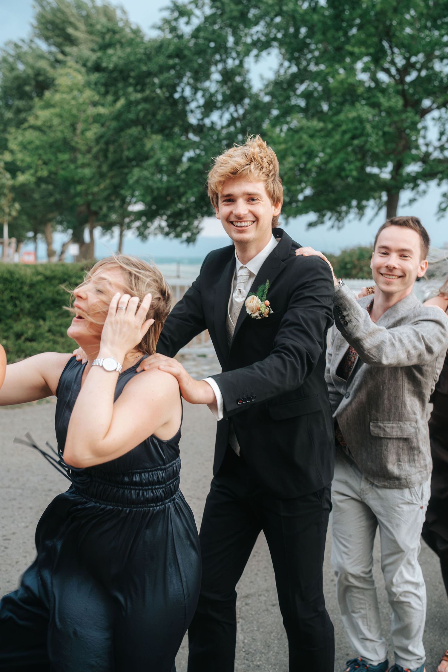 Smiling groom in a black suit with a boutonniere dances outdoors with guests, as a woman in a dark dress laughs beside him.