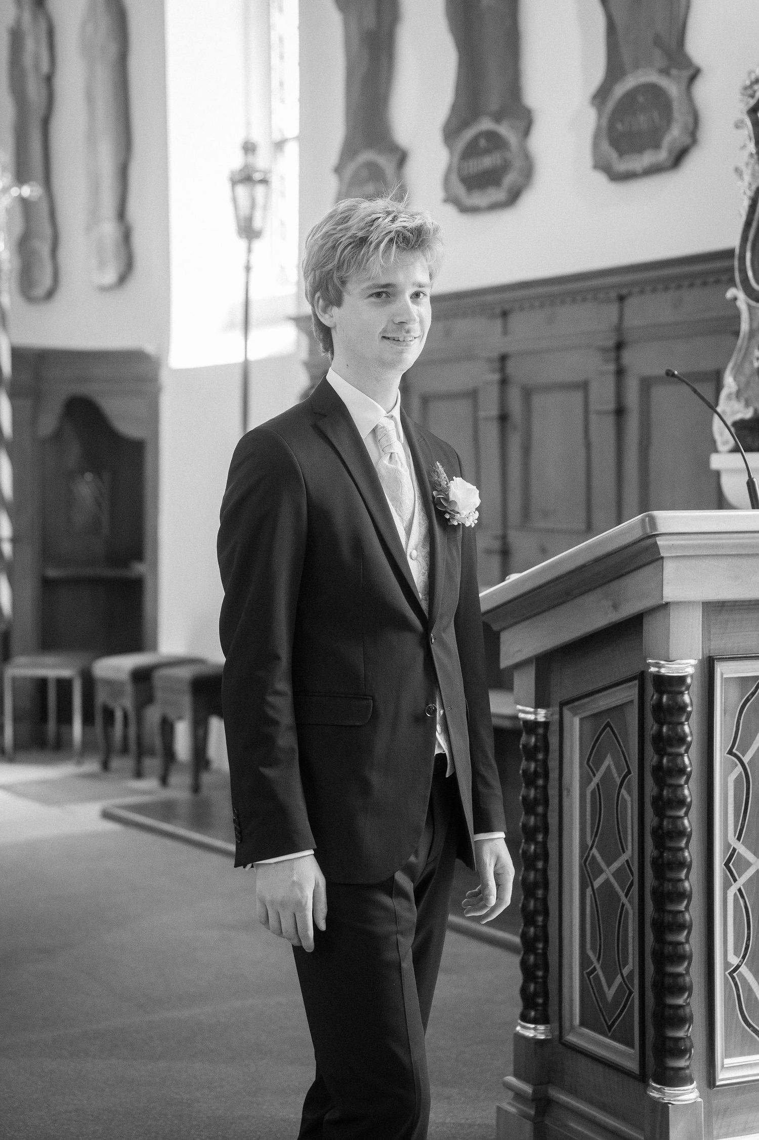 Young groom in black suit with boutonniere stands smiling beside church pulpit on wedding day.
