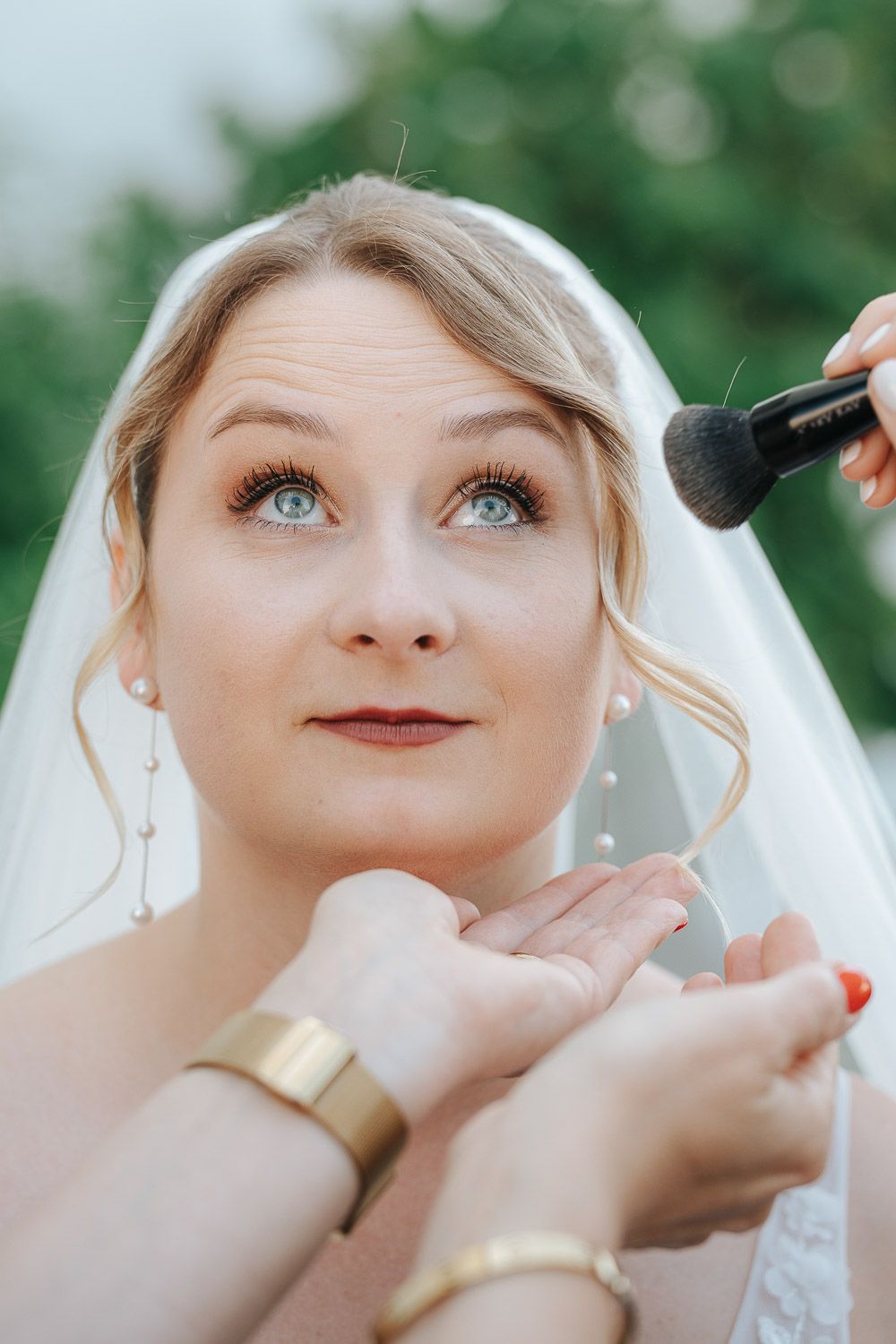A bride getting her makeup done during wedding preparations with a blurry green background.