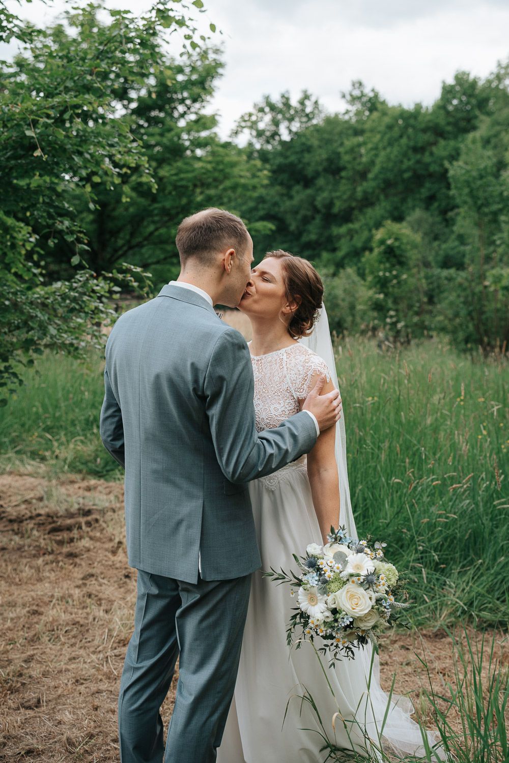 A couple embraces on a tree-lined path during their woodland wedding celebration while holding a white and green bouquet.