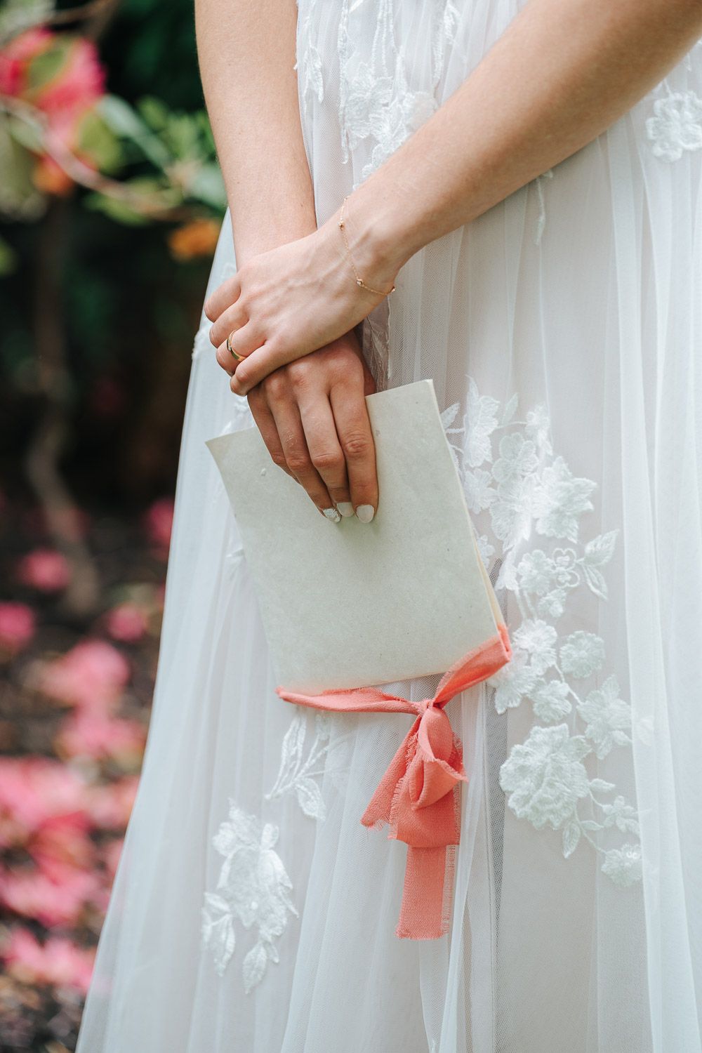 Hands hold a delicate white envelope with coral pink ribbon against a flowing lace wedding dress with pink flowers in background.