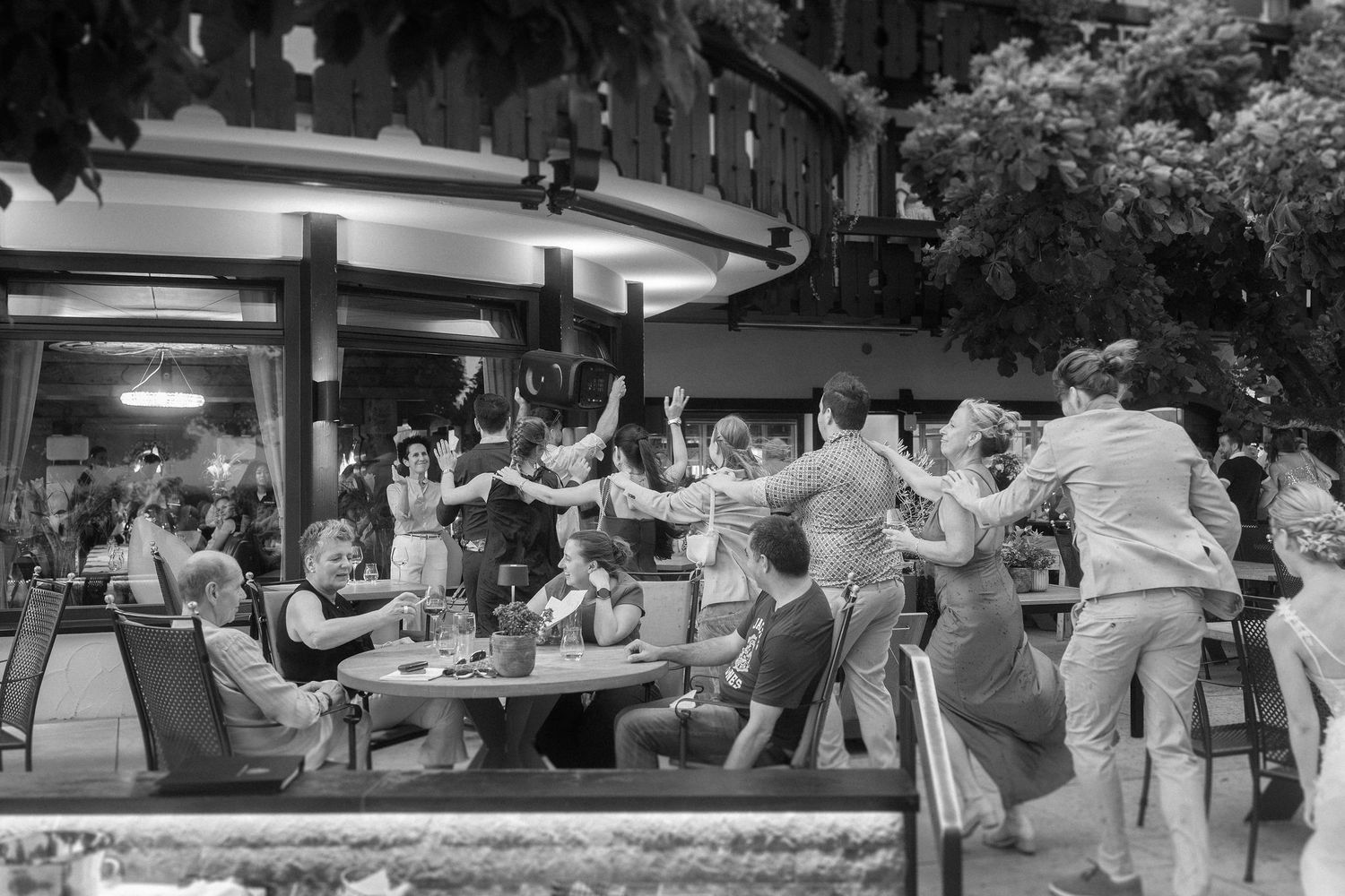 Black and white photo of guests dancing and raising their arms around a table at a lively outdoor patio restaurant party.