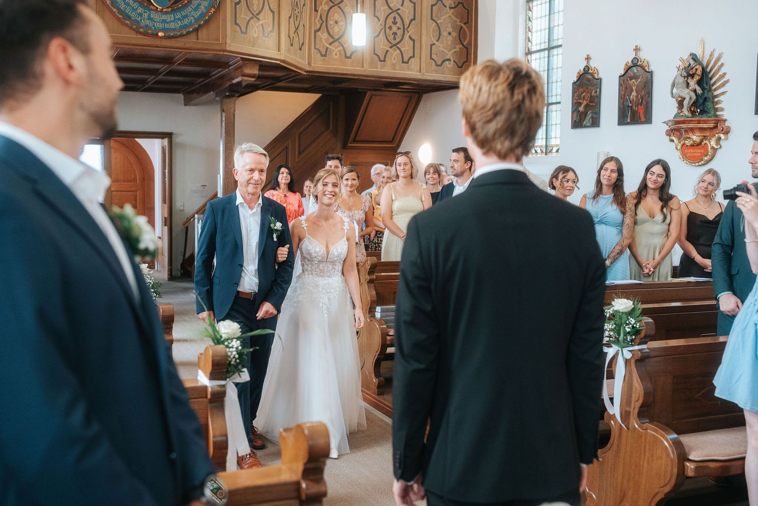 Bride in a white lace gown walks down the church aisle on her father's arm as smiling wedding guests in pews watch the ceremony.