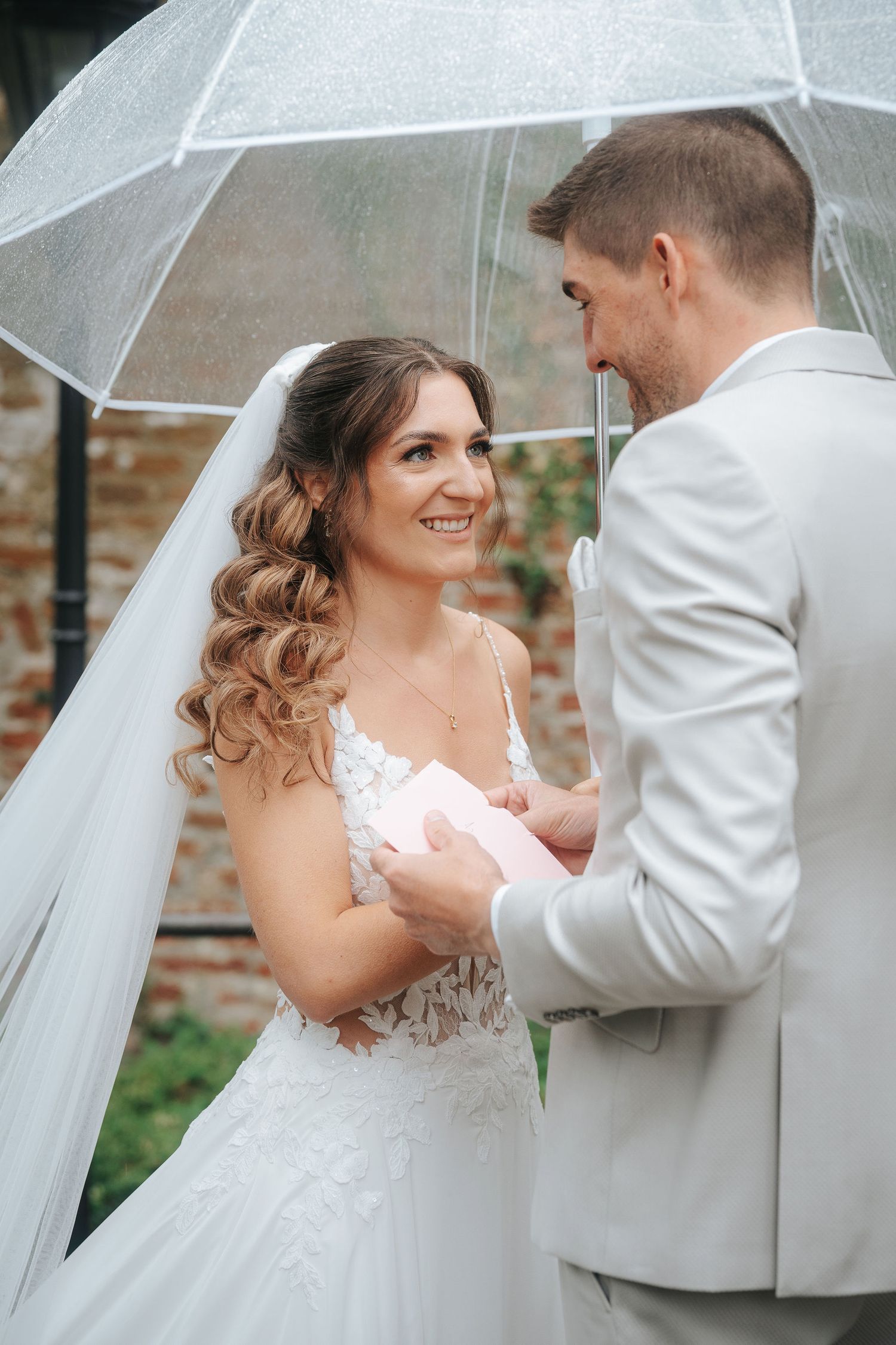 Smiling bride in a lace wedding dress and veil stands under a clear umbrella as the groom in a light gray suit reads her his vows.