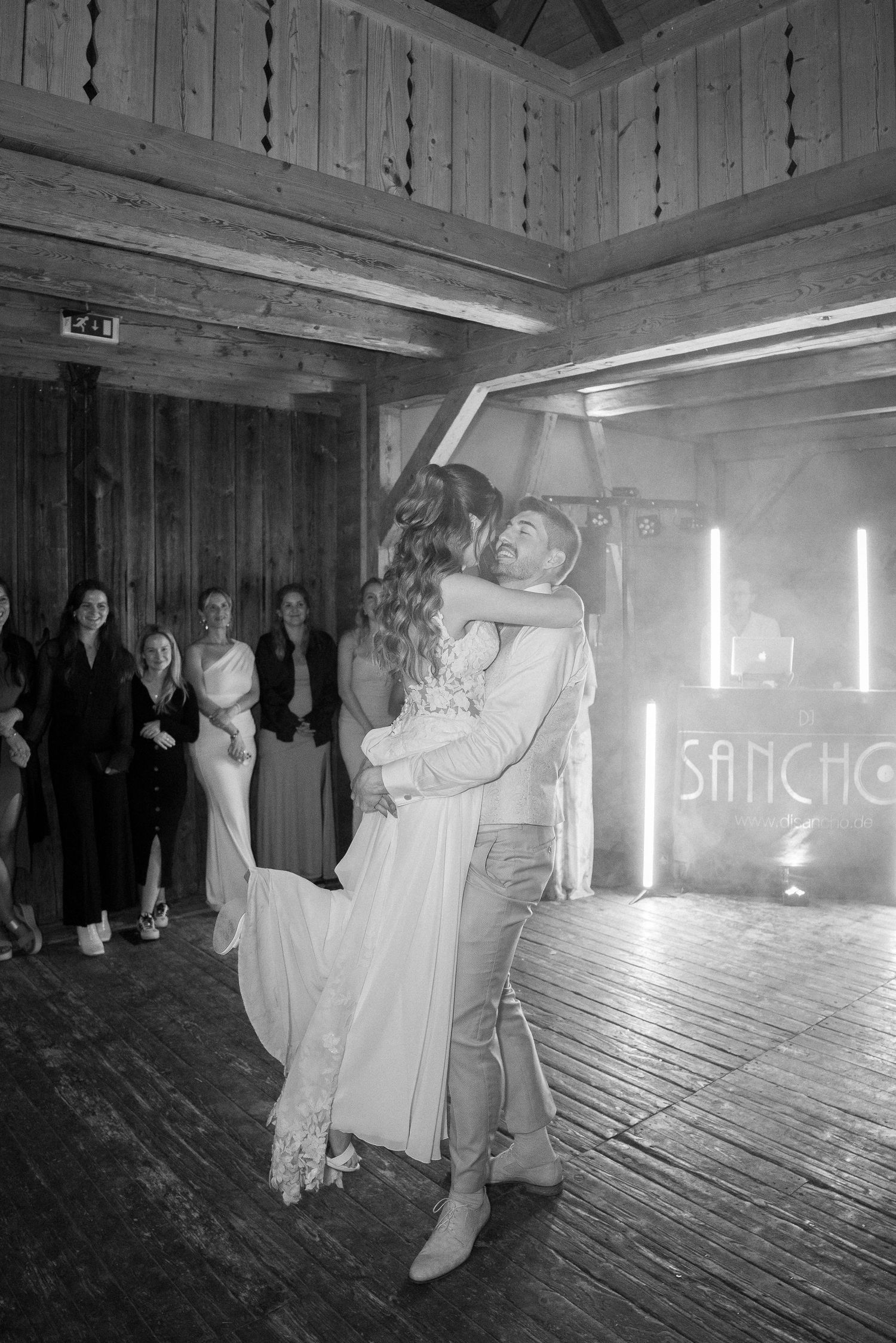 Bride and groom embrace in a first dance on a rustic wooden barn floor while wedding guests watch in black and white.