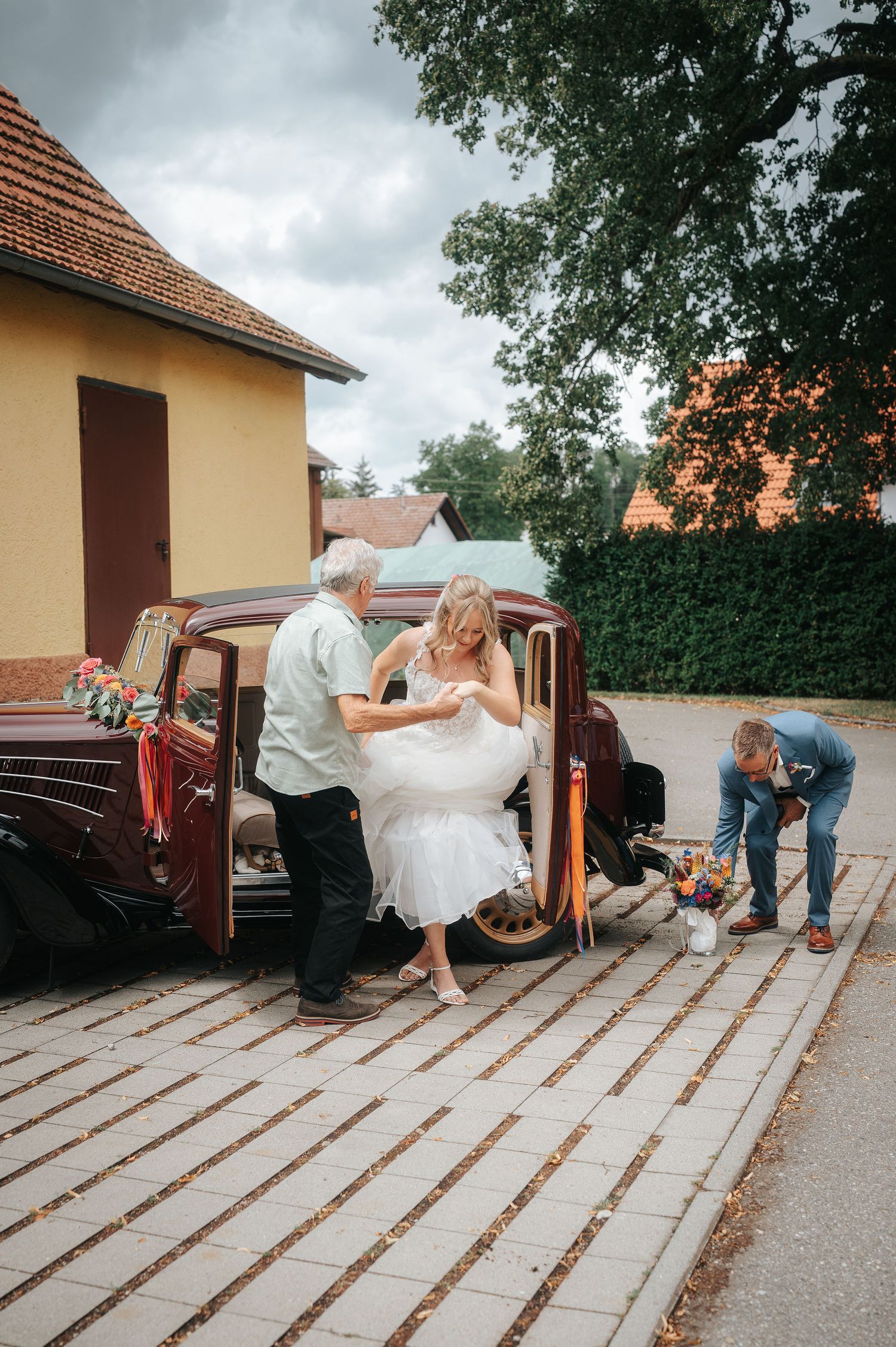 Bride in a white wedding gown steps out of a vintage car as an older man helps her, while a groom in a blue suit kneels nearby.