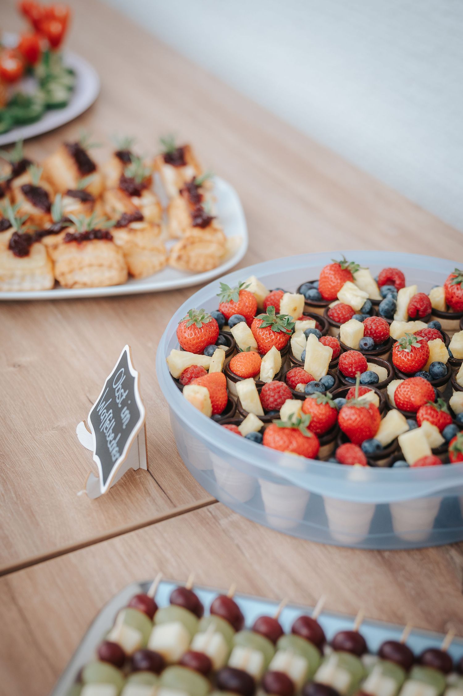 Buffet table with a fruit dessert cup topped with strawberries, blueberries and cheese cubes, plus canapés and skewers in the background.