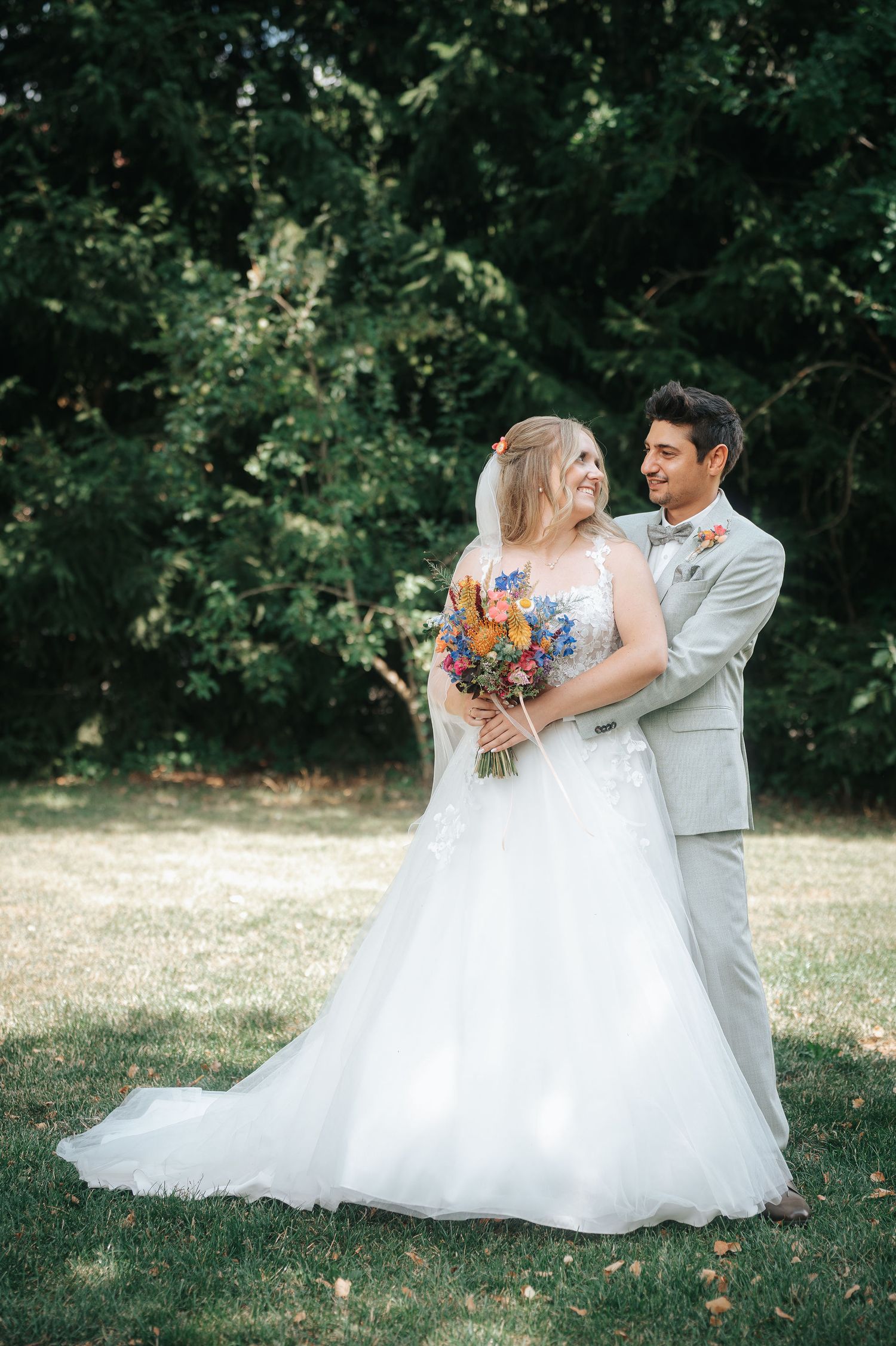 Smiling bride in a white lace gown holding a colorful wildflower bouquet is embraced by her groom in a gray suit on a sunny lawn.