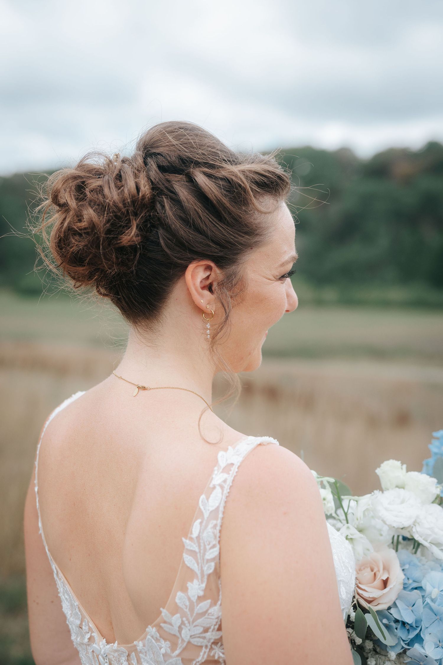 Bride in a lace open-back wedding dress with a braided updo holds a blue and white floral bouquet while looking out over a grassy field.