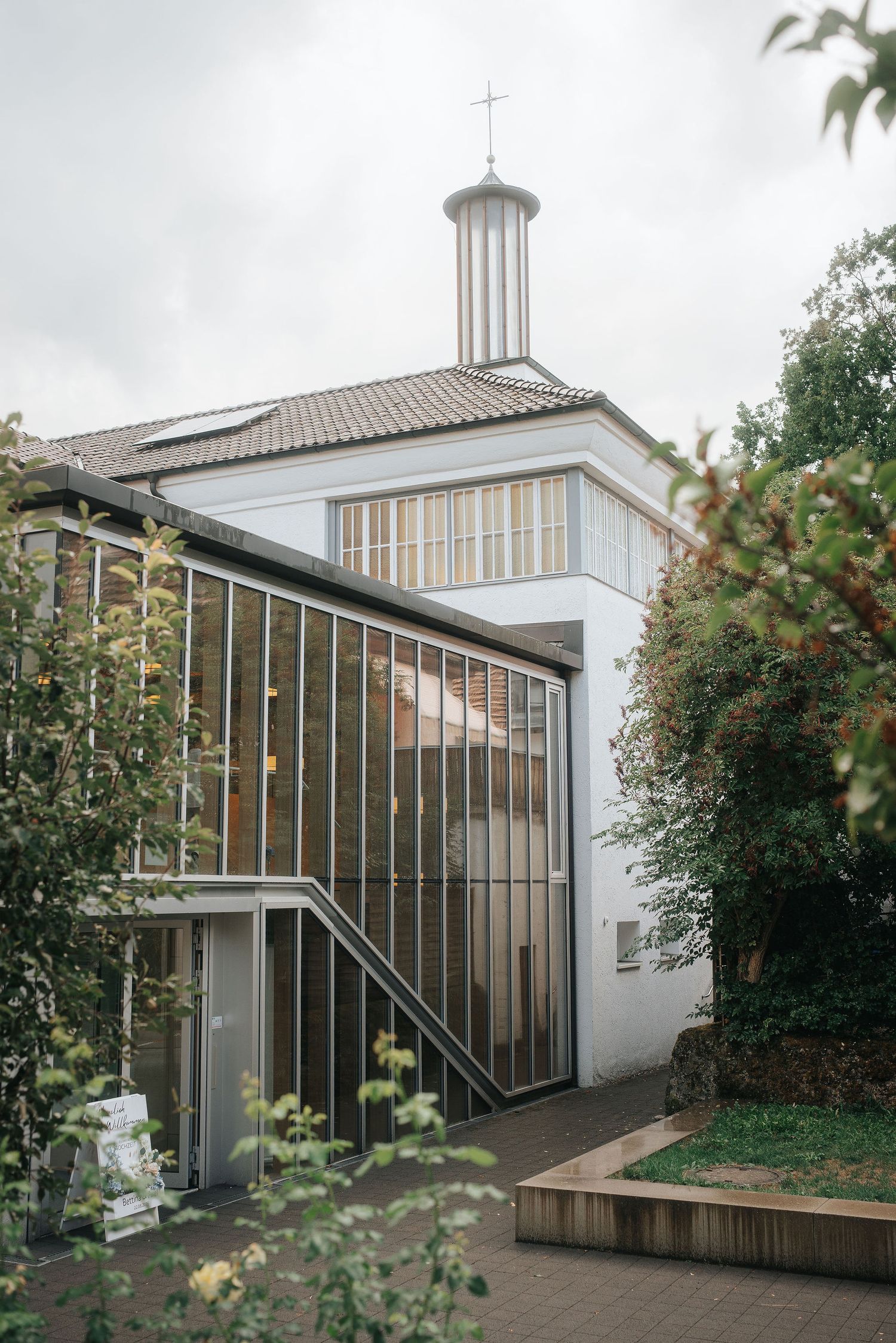 Modern church building with a glass-and-steel entrance, white walls, tiled roof, and a slender cross-topped tower, framed by green trees.