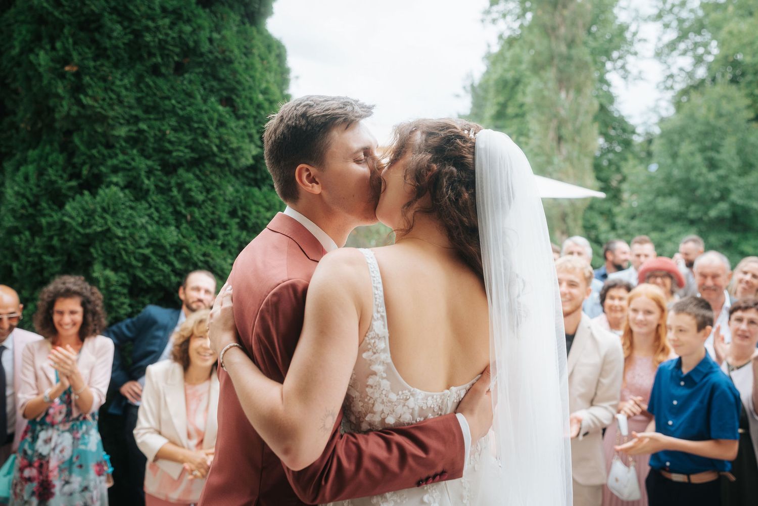 Bride in a lace gown and veil kisses her groom in a rust-colored suit while smiling wedding guests watch outdoors.