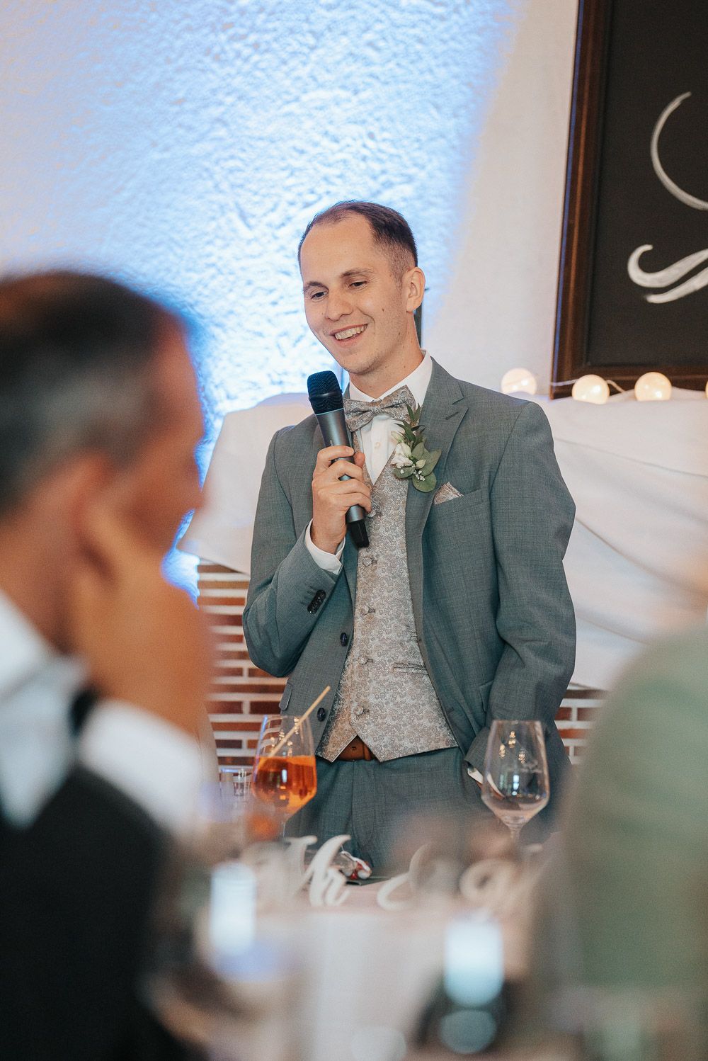 A wedding guest in a gray suit and bow tie gives a speech while holding a microphone at an elegant reception.