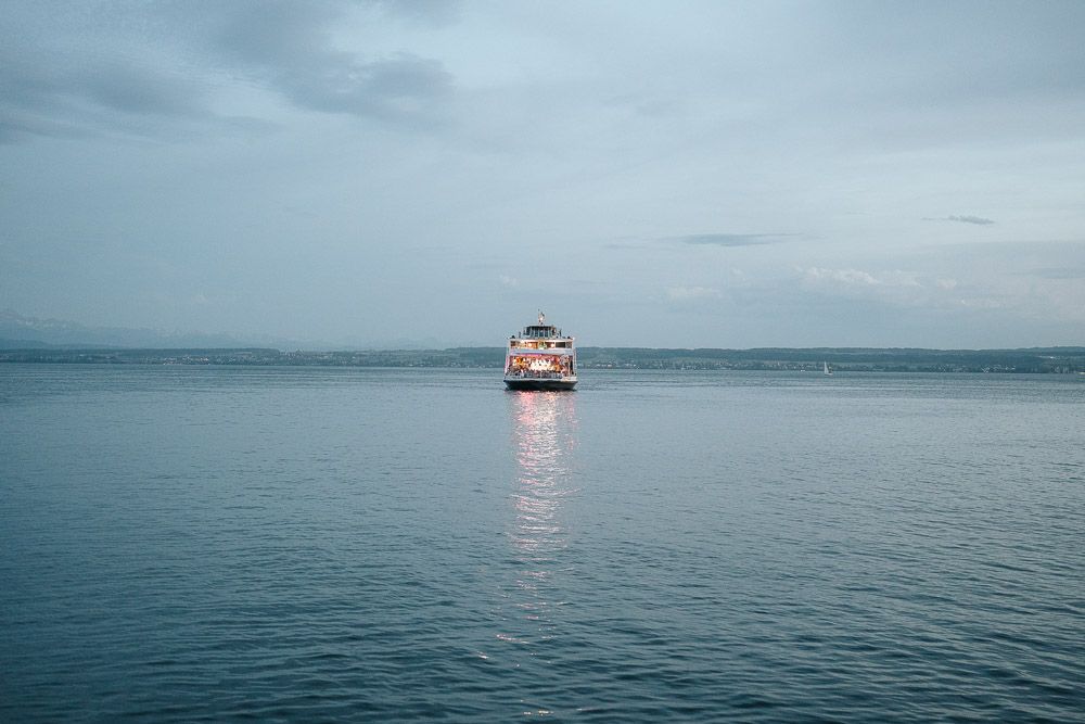 A lone ferry boat crosses calm waters under an overcast sky during twilight, casting reflections on the misty seascape.