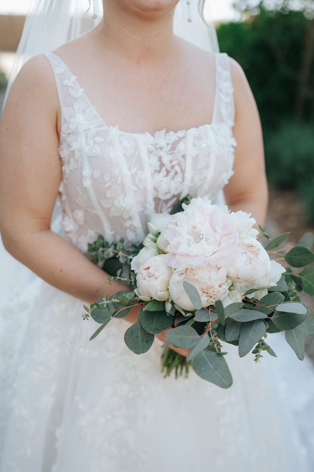 A delicate bridal bouquet of blush pink roses and eucalyptus leaves held against a white lace wedding dress.