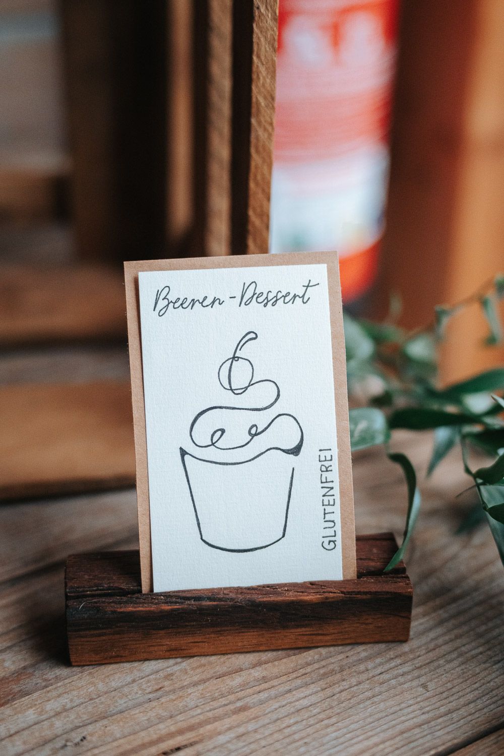 A simple hand-drawn sketch of a cupcake on a card labeled 'Beeren-Dessert' displayed in a wooden holder.