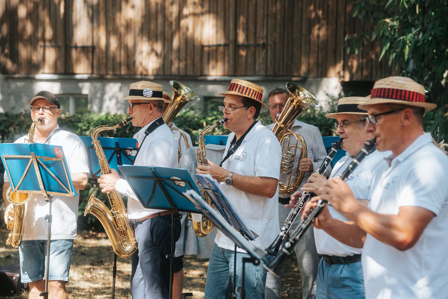 Brass band musicians in white shirts and straw hats play saxophones and trumpets outdoors from sheet music on stands.